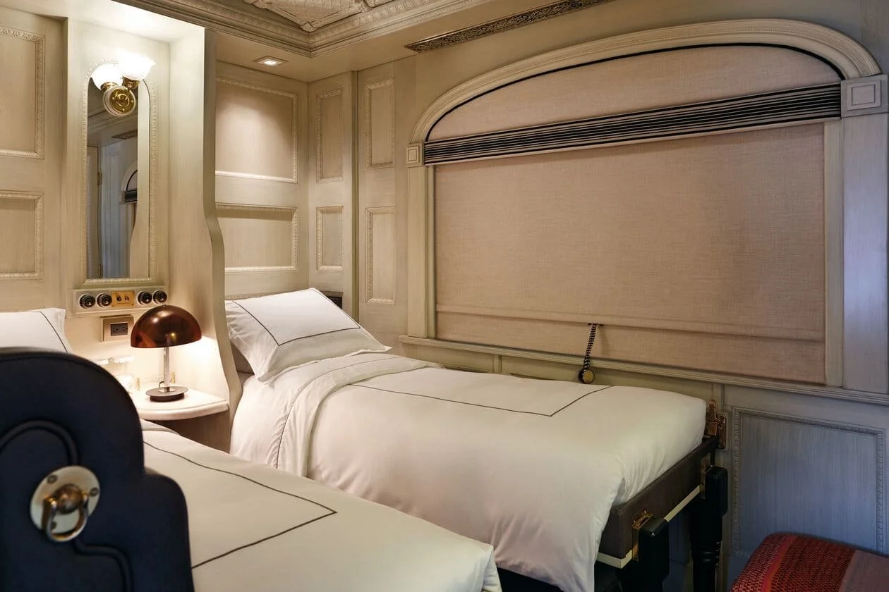Hotel room with two single beds, ornate wall paneling, a mirror, a small bedside lamp, and a window covered by a beige curtain.