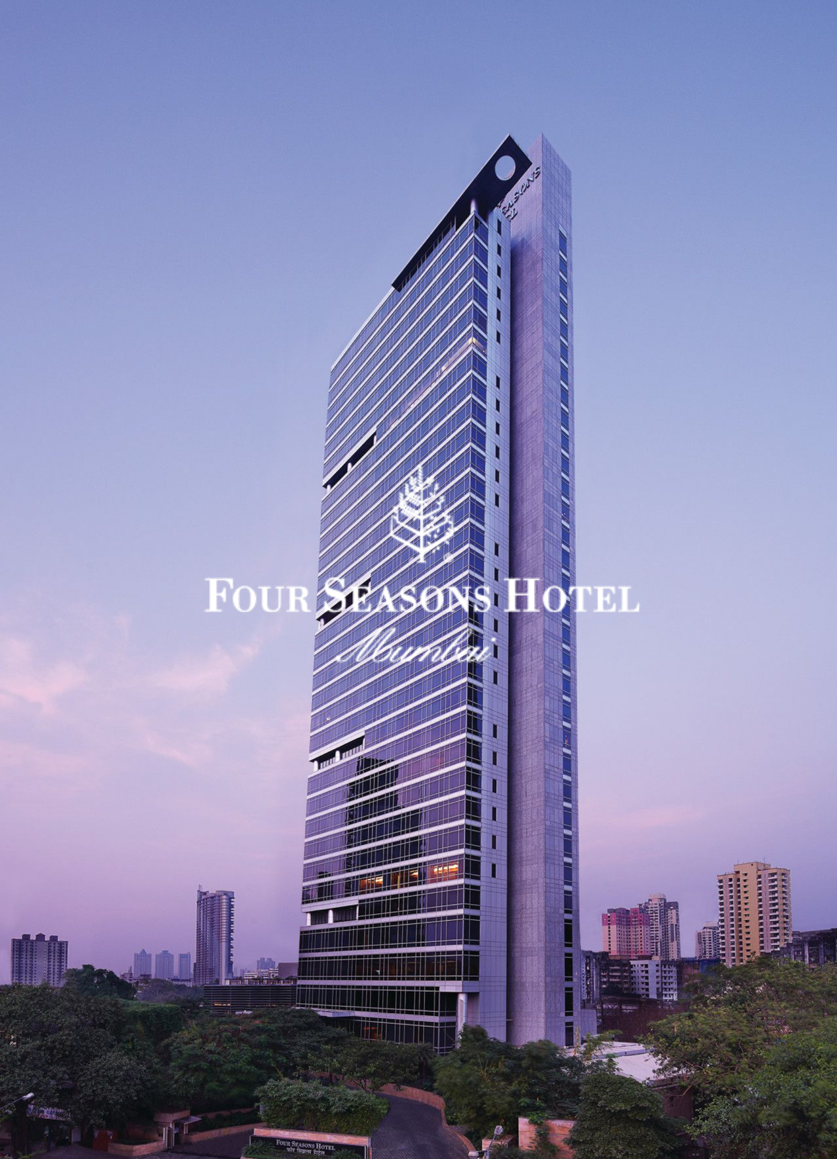 Tall modern hotel building with glass facade and the Four Seasons logo at the top, city skyline visible in the background, and trees in the foreground during dusk.