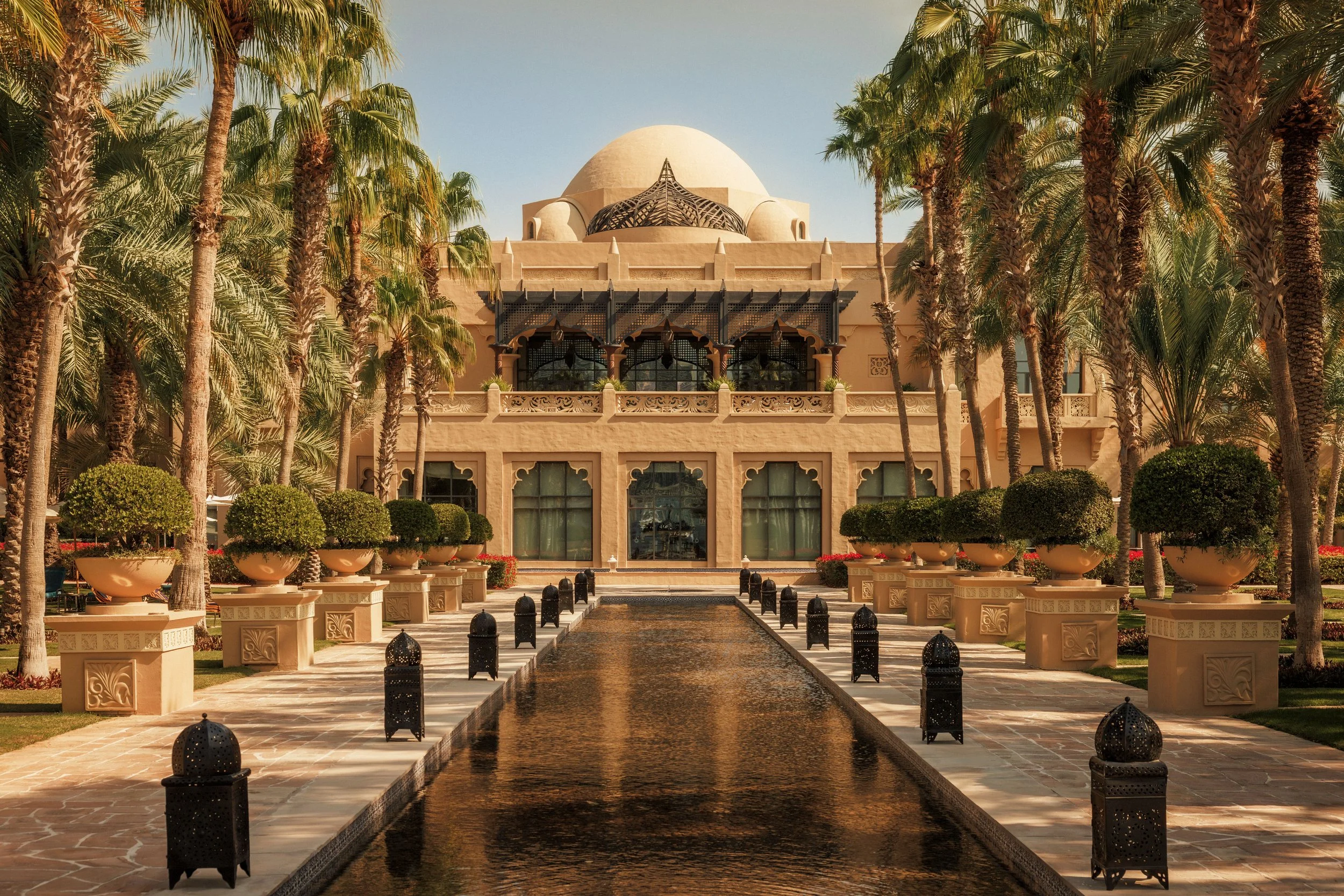 A luxurious desert-style building with a domed roof, surrounded by palm trees, potted plants, and a water feature with lanterns lining a pathway.