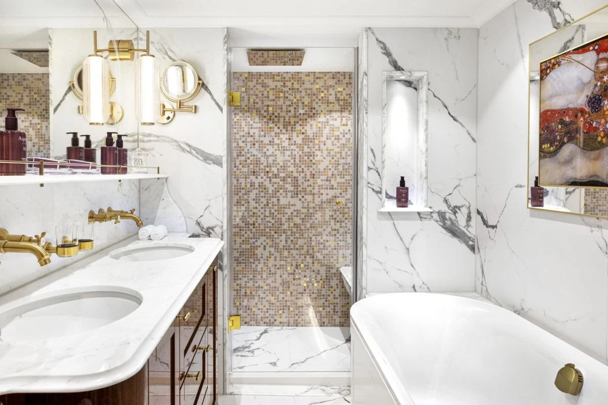 Luxurious bathroom with marble walls and floor, featuring a double vanity with gold fixtures, bottles of soap or lotion, a large bathtub, a shower with a mosaic tile wall, a small window, and a framed colorful painting.