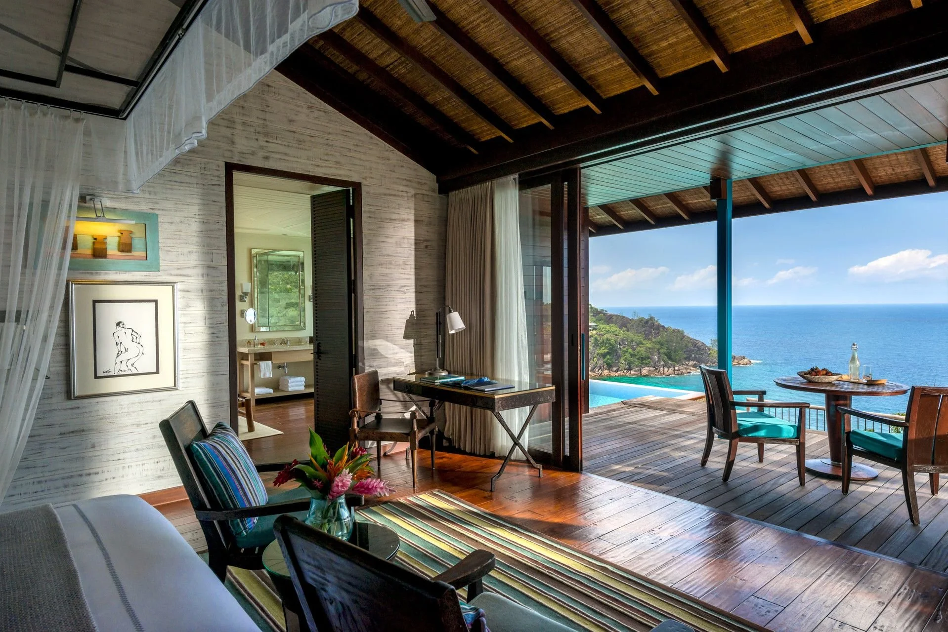 Interior view of a beachside room with an open sliding door leading to a balcony with a view of the ocean, green hills, and rocky coastline. The room has a wooden ceiling, light-colored textured walls, and wooden flooring. There are chairs with striped and solid cushions, a small table with flowers, and artwork on the walls.
