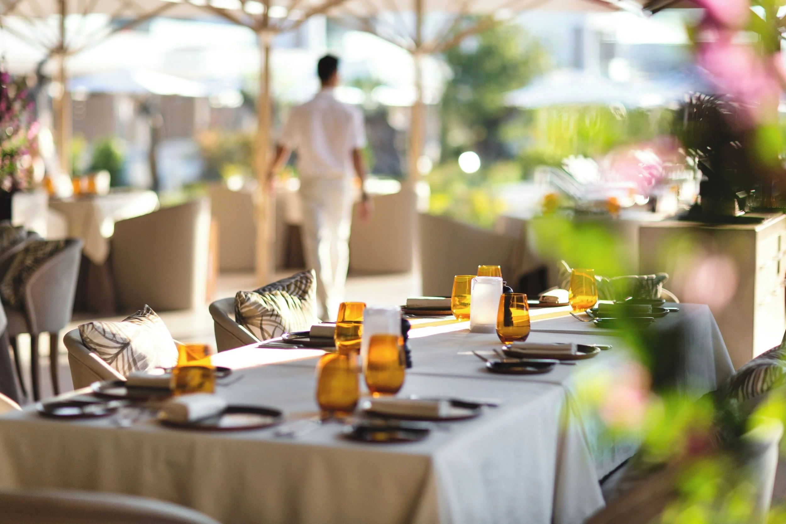 A blurred image of an outdoor dining area with a table set with orange glasses, white plates, and napkins. In the background, there are umbrellas and a person standing.