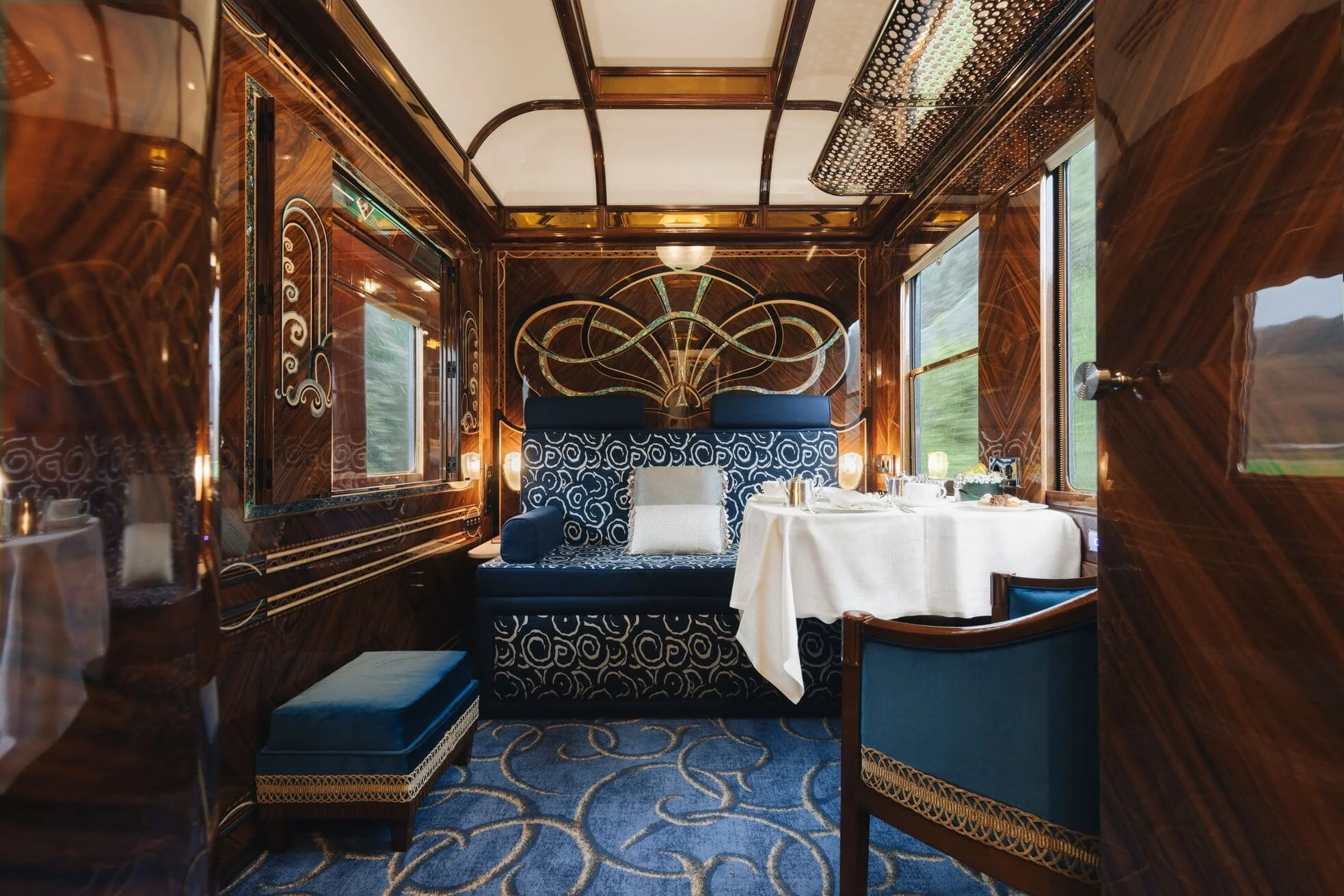 Interior of a luxurious dining car with polished wood paneling, ornate decoration, a booth with blue patterned upholstery, and a table set for a meal near large windows showing a blurred outdoor landscape.