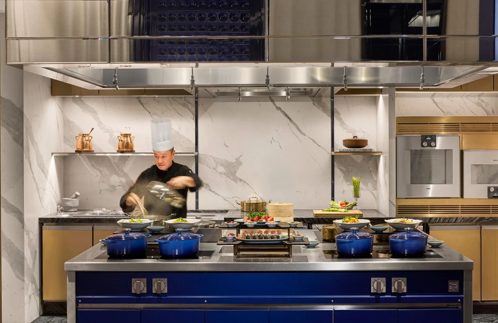A chef preparing dishes in a modern, upscale kitchen with a marble backsplash and a variety of plated food on a countertop.