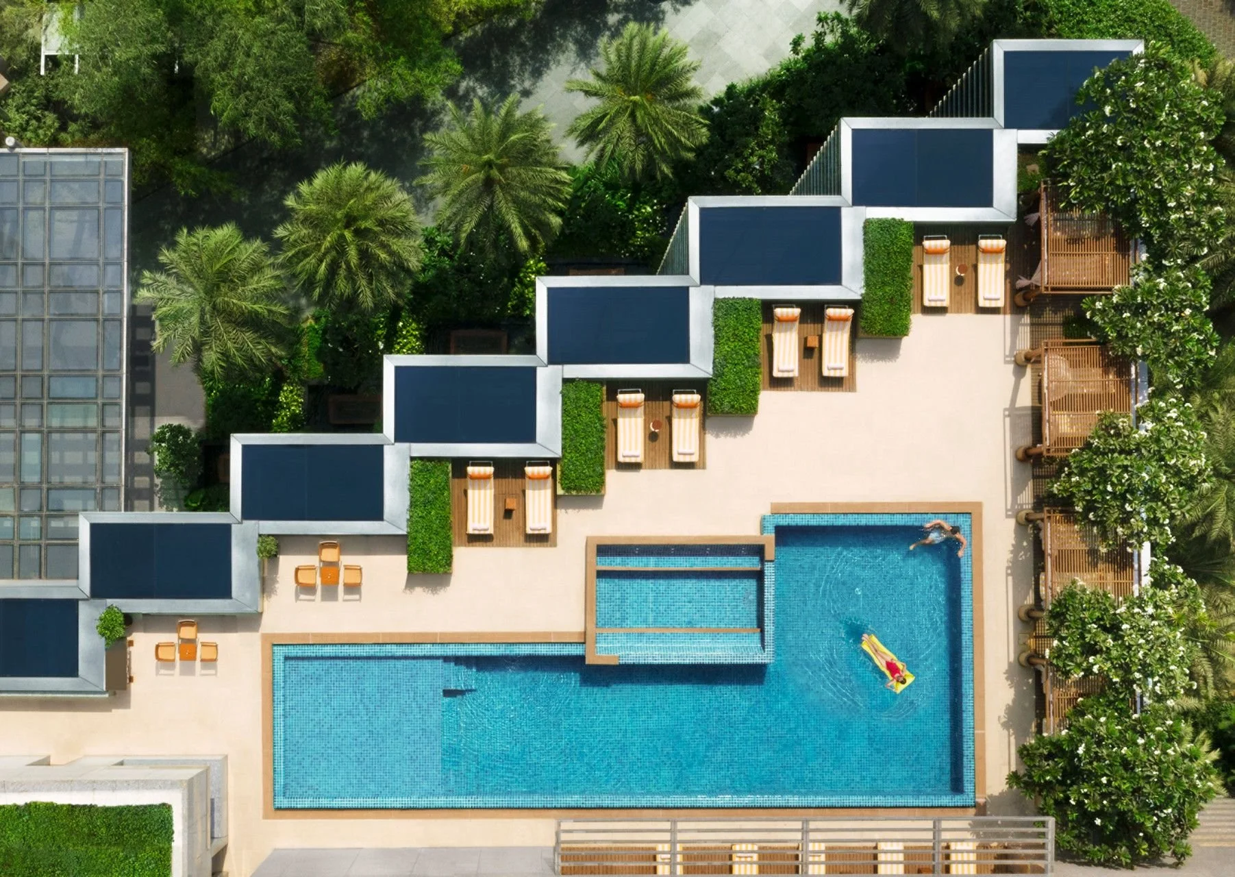 Aerial view of a swimming pool area with lounge chairs, sitting areas under wooden pergolas, surrounded by lush green trees and plants, adjacent to modern buildings.