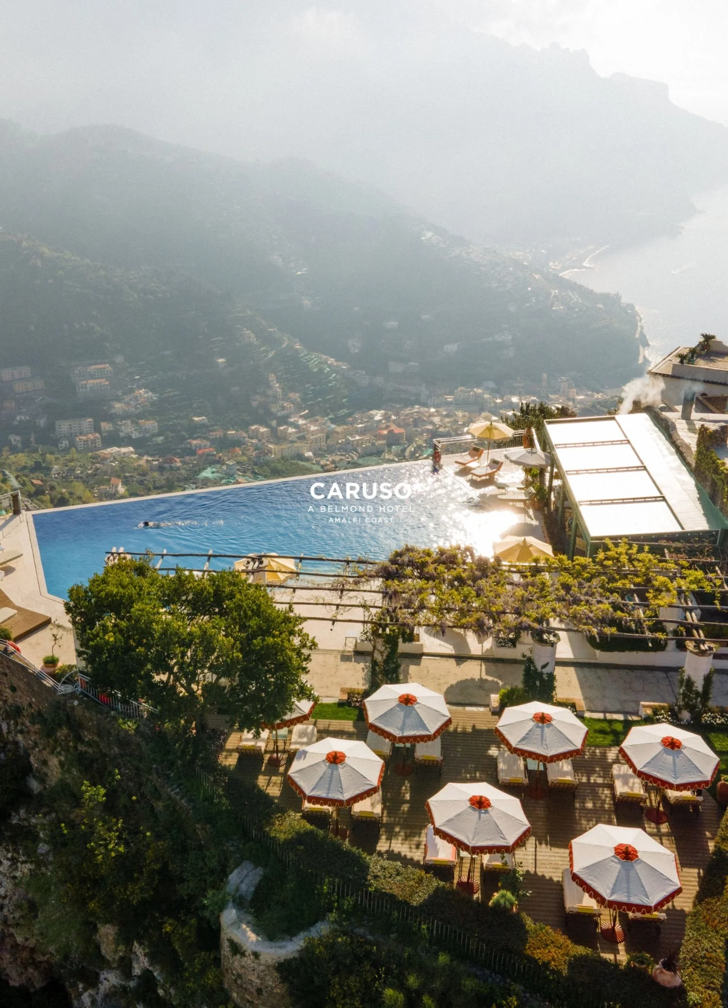 An infinity pool overlooking a city and coastline with umbrellas and lounge chairs around the pool area at Caruso, a Belmond Hotel, Amalfi Coast.