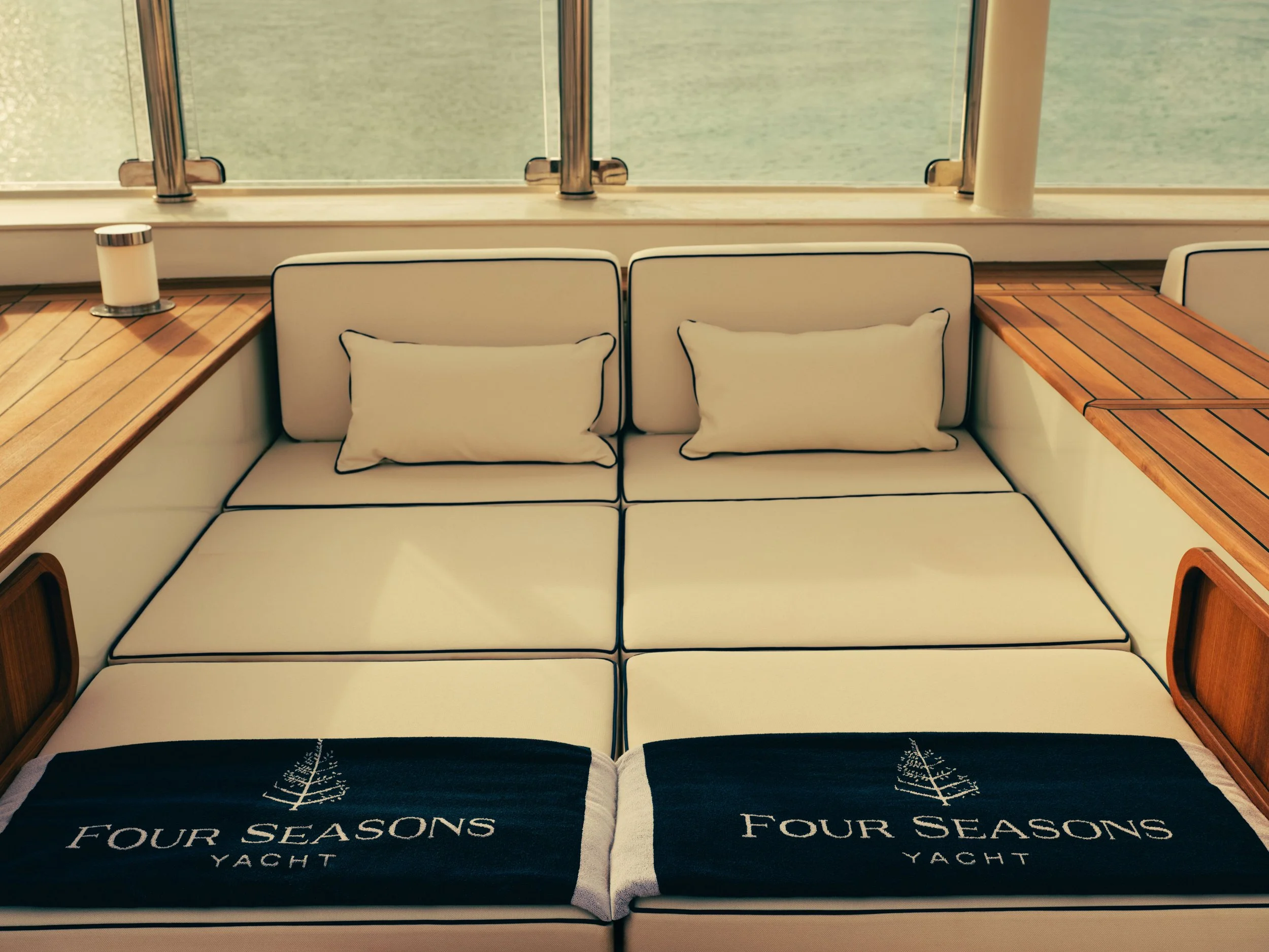 A sofa with white cushions and black outline, with a black blanket featuring white trees and the text 'FOUR SEASONS YACHT' in front, next to a wooden table with a candle in a glass holder, set near a large window with a view of water outside.