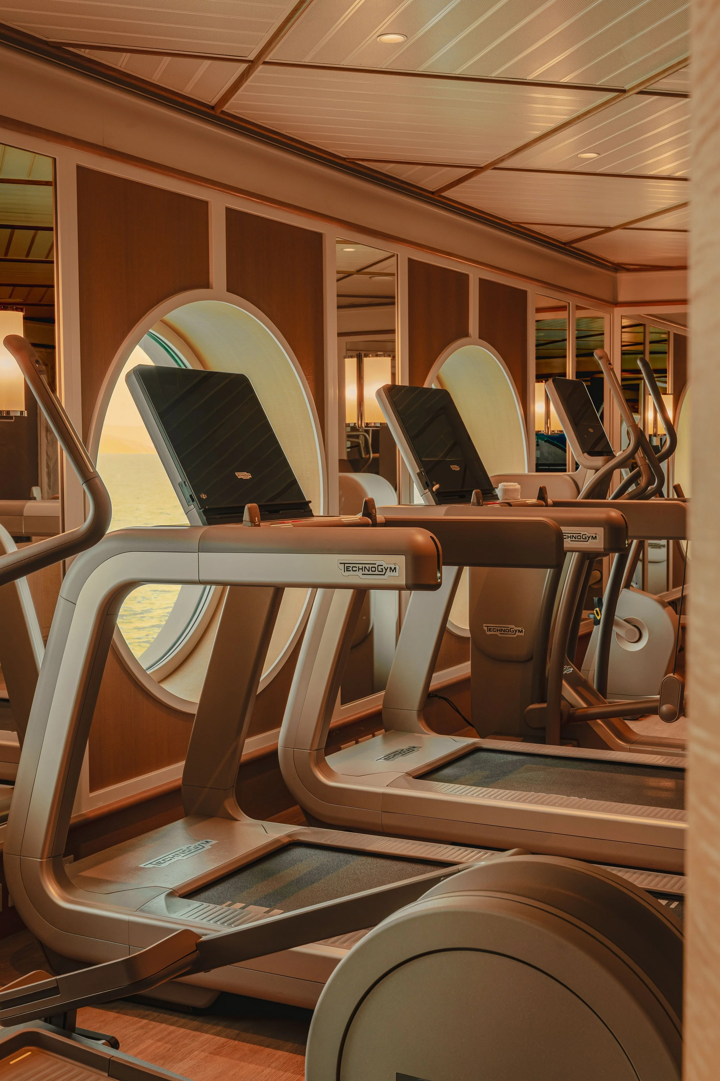 Row of exercise treadmills in a gym with large windows and wooden panel walls.