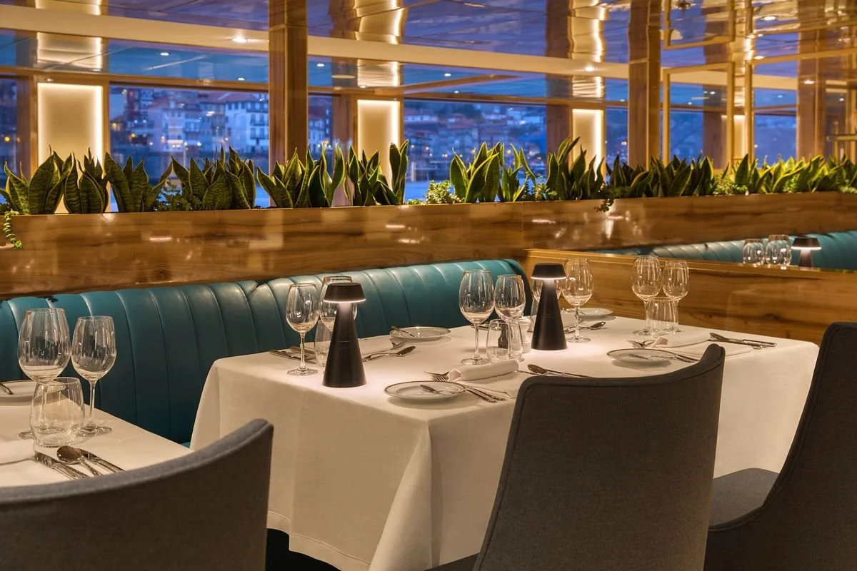 Interior of a restaurant with dining tables set with white tablecloths, wine glasses, plates, cutlery, black lamps, and green upholstered chairs, with a lush green plant divider and large windows showing city lights at night.