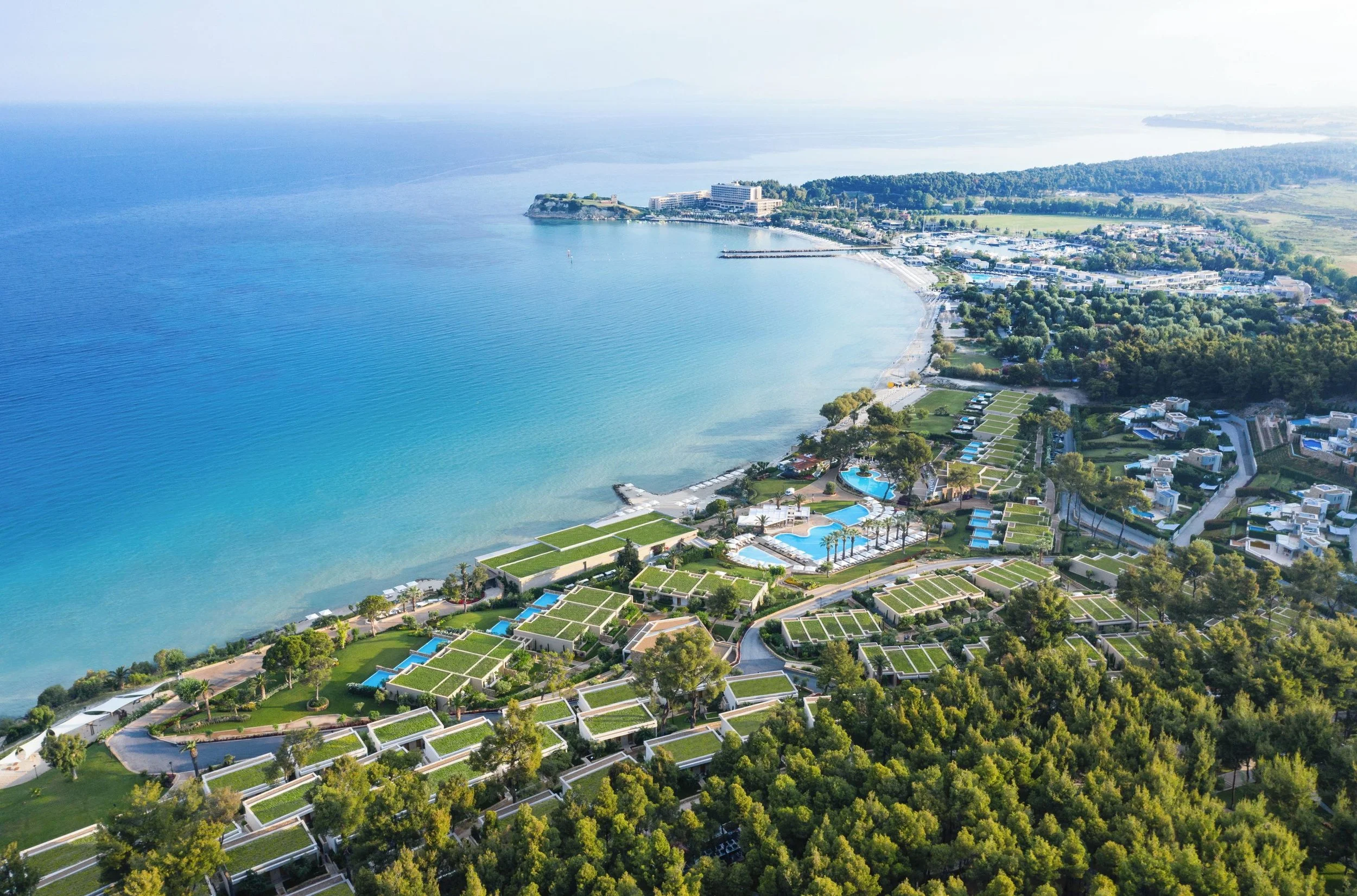 Aerial view of a coastal resort area with a curved shoreline, clear blue water, white buildings with green roofs, swimming pools, and lush greenery.