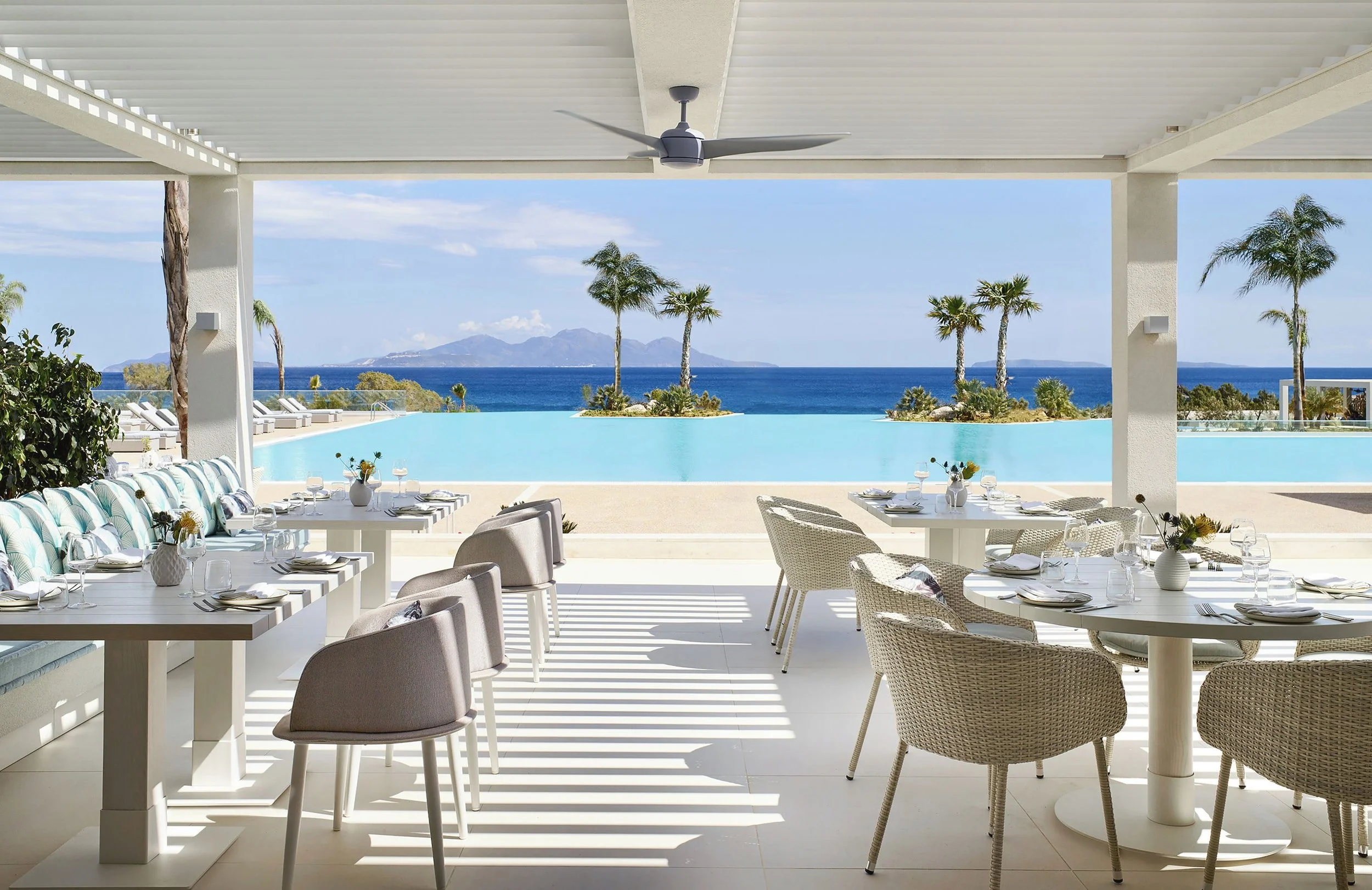Outdoor dining area with white tables and chairs overlooking a pool, palm trees, ocean, and mountains in the background.