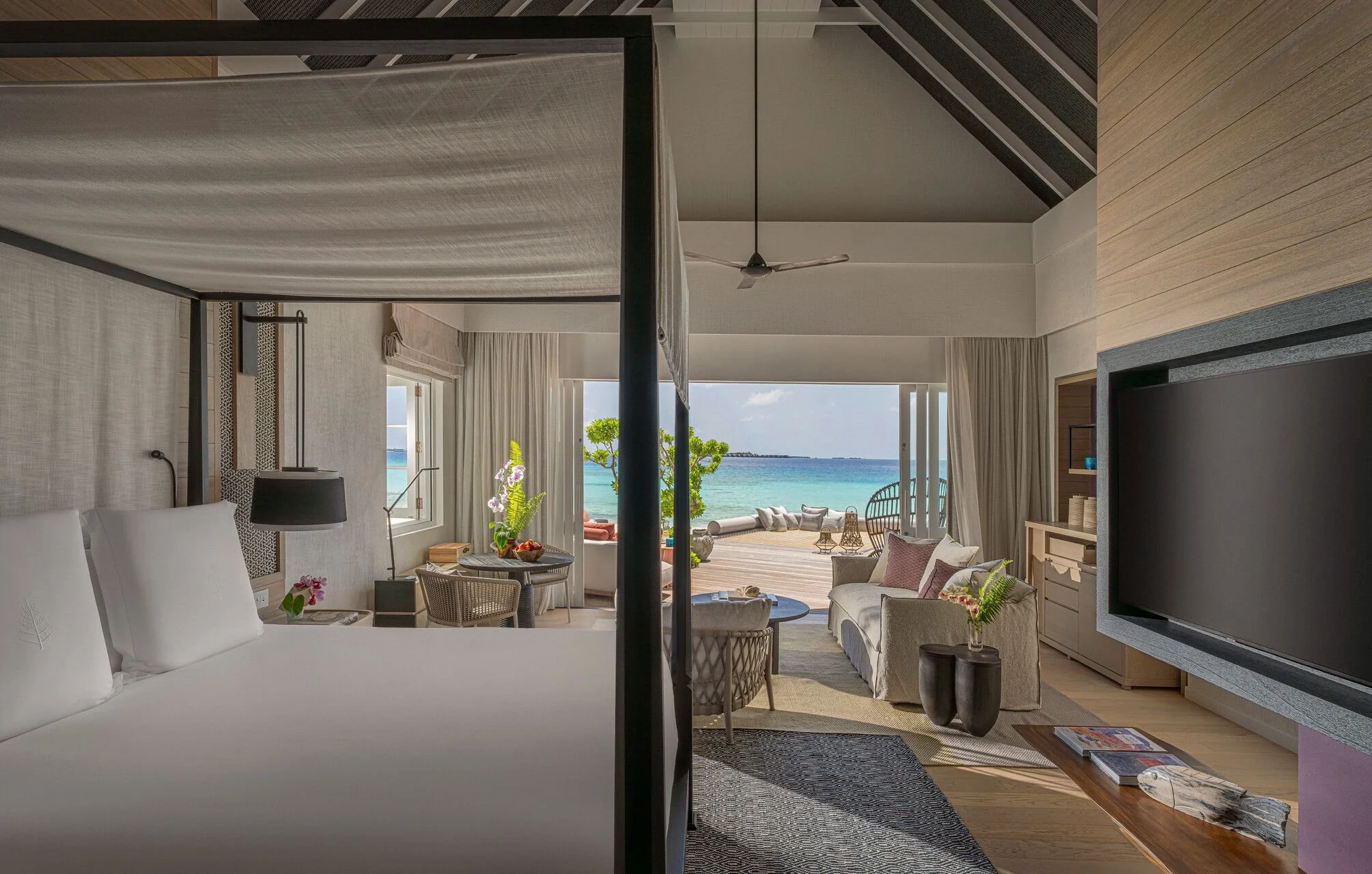 A hotel room with a view of the beach and ocean, featuring a four-poster bed, seating area with cream-colored sofas, and large windows with curtains.