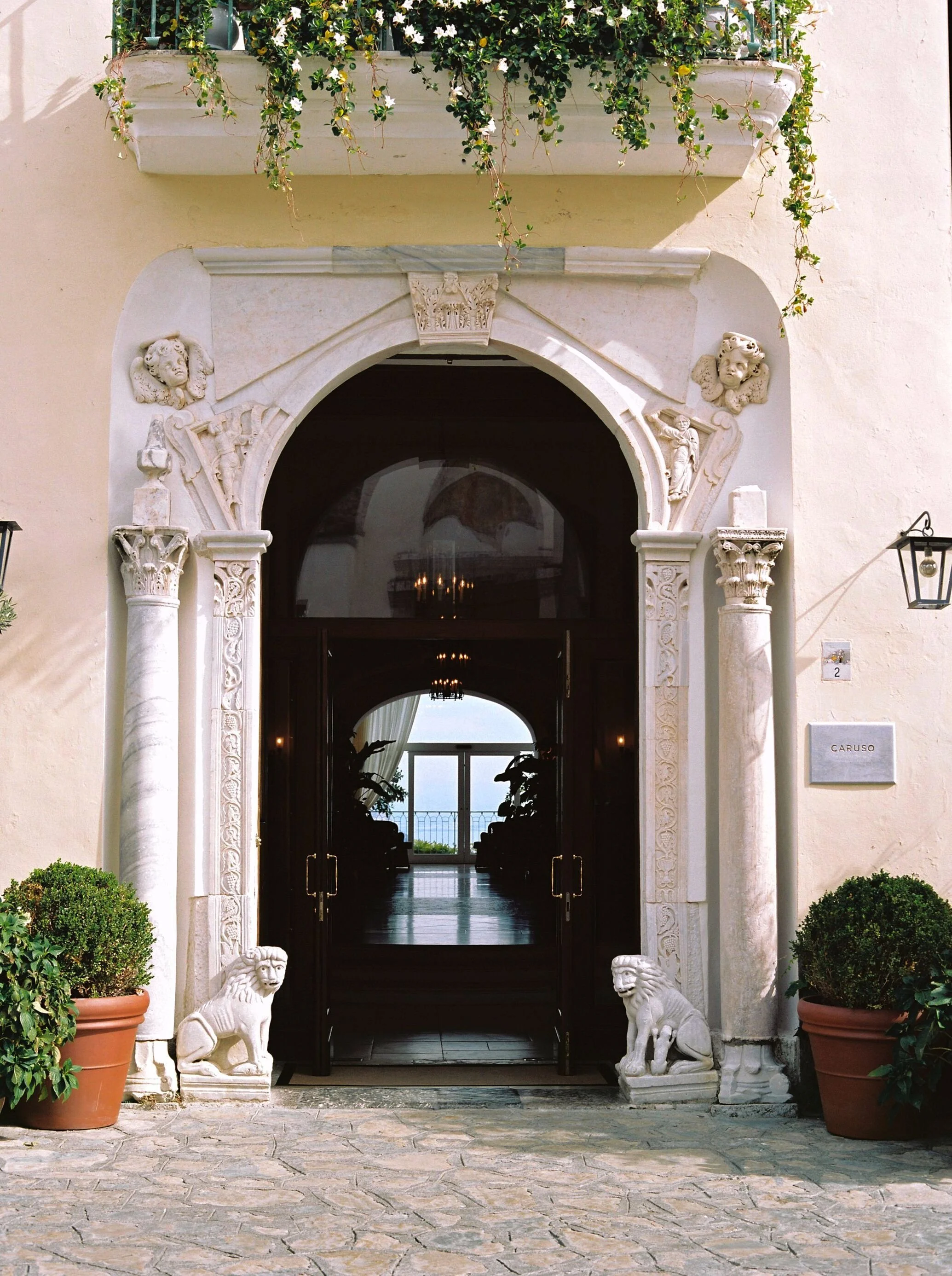 The entrance to a building with neoclassical architecture, featuring an ornate archway supported by two decorated columns. Two white lion statues flank the doorway, and there are potted plants on either side. The door opens into a room with a view of the sea and a chandelier hanging inside.