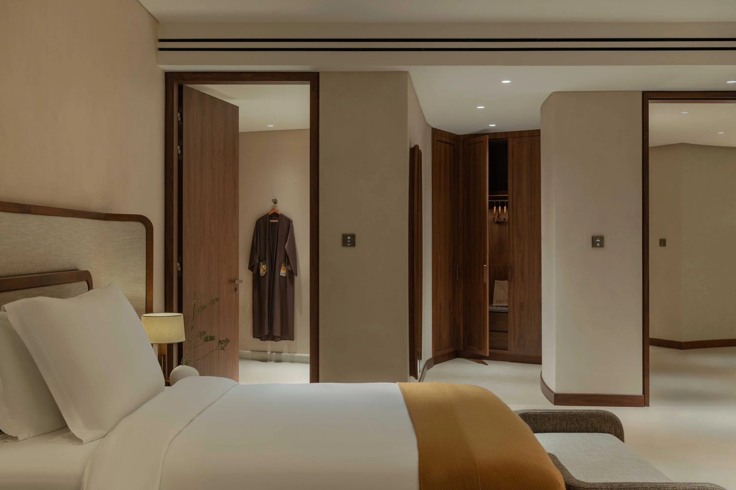 A bedroom with a bed, a beige headboard, and white bedding. There is a bedside lamp and a small plant. An open door reveals a walk-in closet with wooden shelving. The room has beige walls, wooden trim, and recessed ceiling lights.