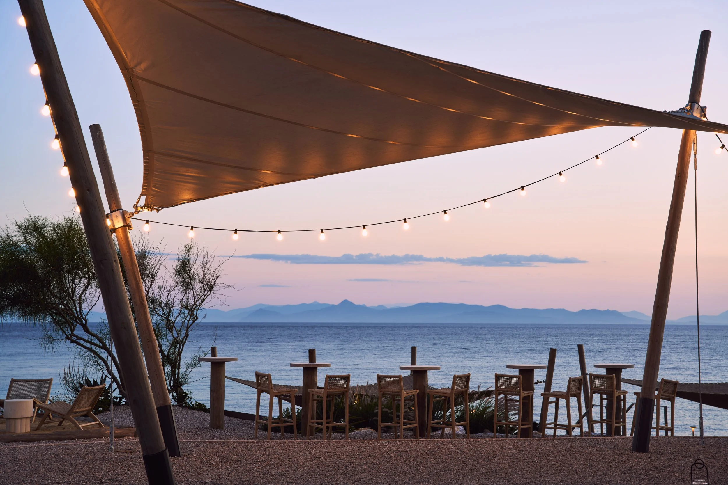 Beachside outdoor seating area with chairs and tables, large fabric shade sail, string lights, and a view of the water with mountains in the distance at dusk.