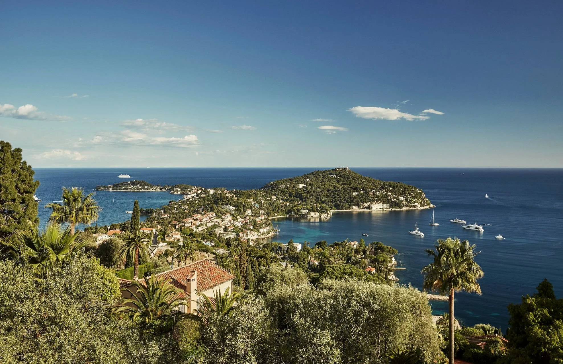 A coastal town with lush greenery, palm trees, and a hillside overlooking the ocean. There are boats and ships in the water, and buildings along the coastline under a clear blue sky.