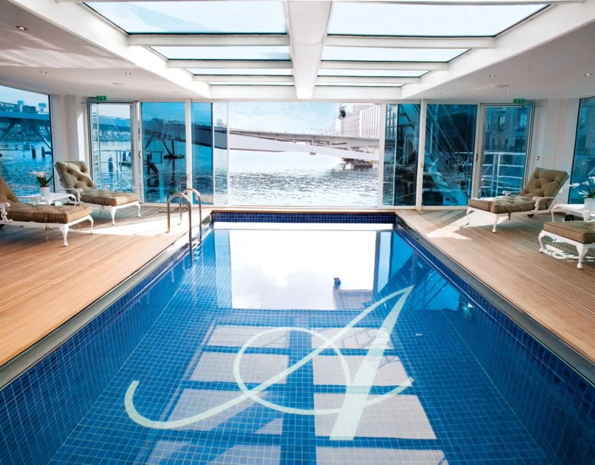 Indoor swimming pool with city and waterview outside large glass windows, four lounge chairs, and wooden floor.
