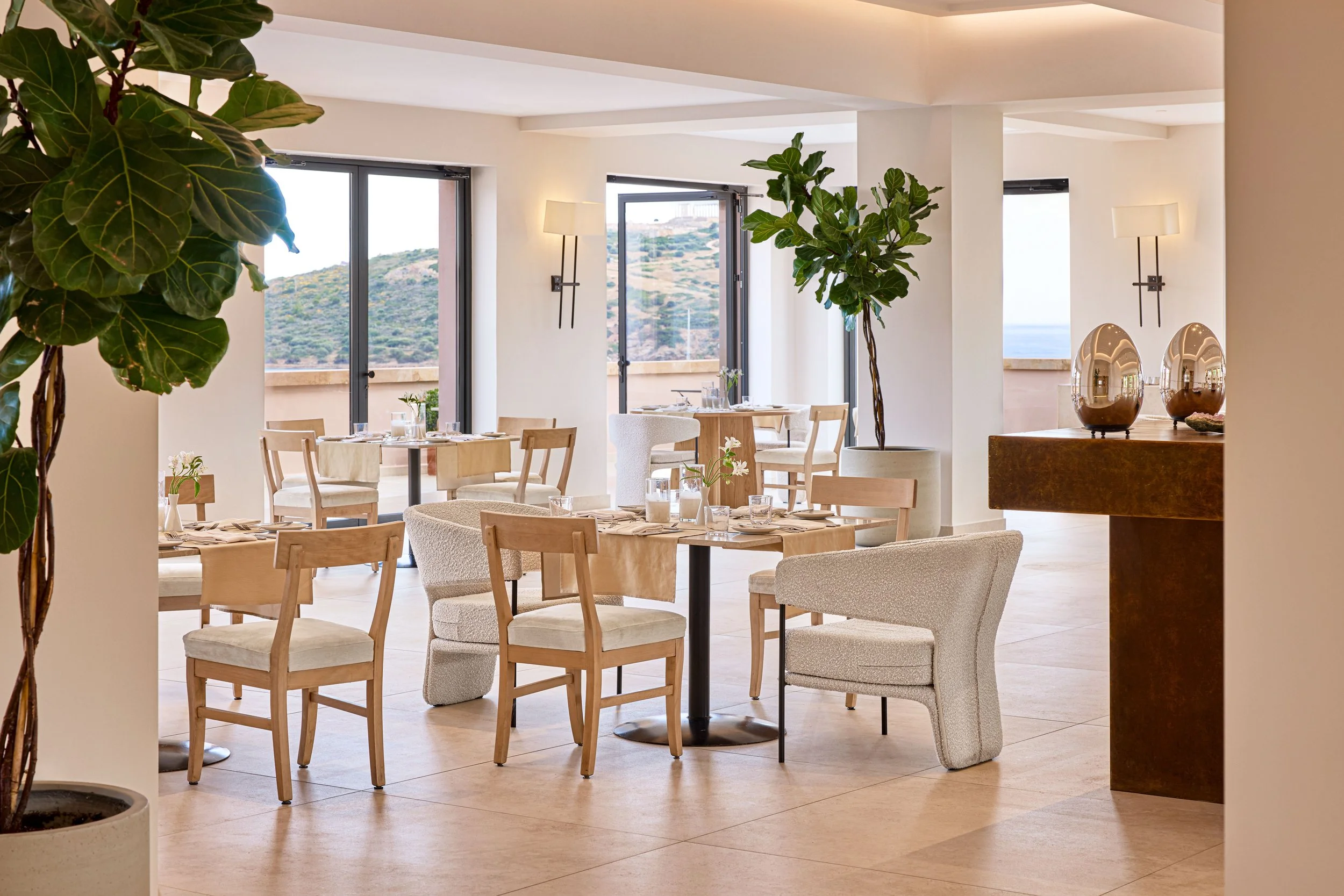 Bright, modern dining area with round wooden tables, cream upholstered chairs, large windows with scenic hillside view, and minimalistic decor including two decorative vases on a wooden side table.