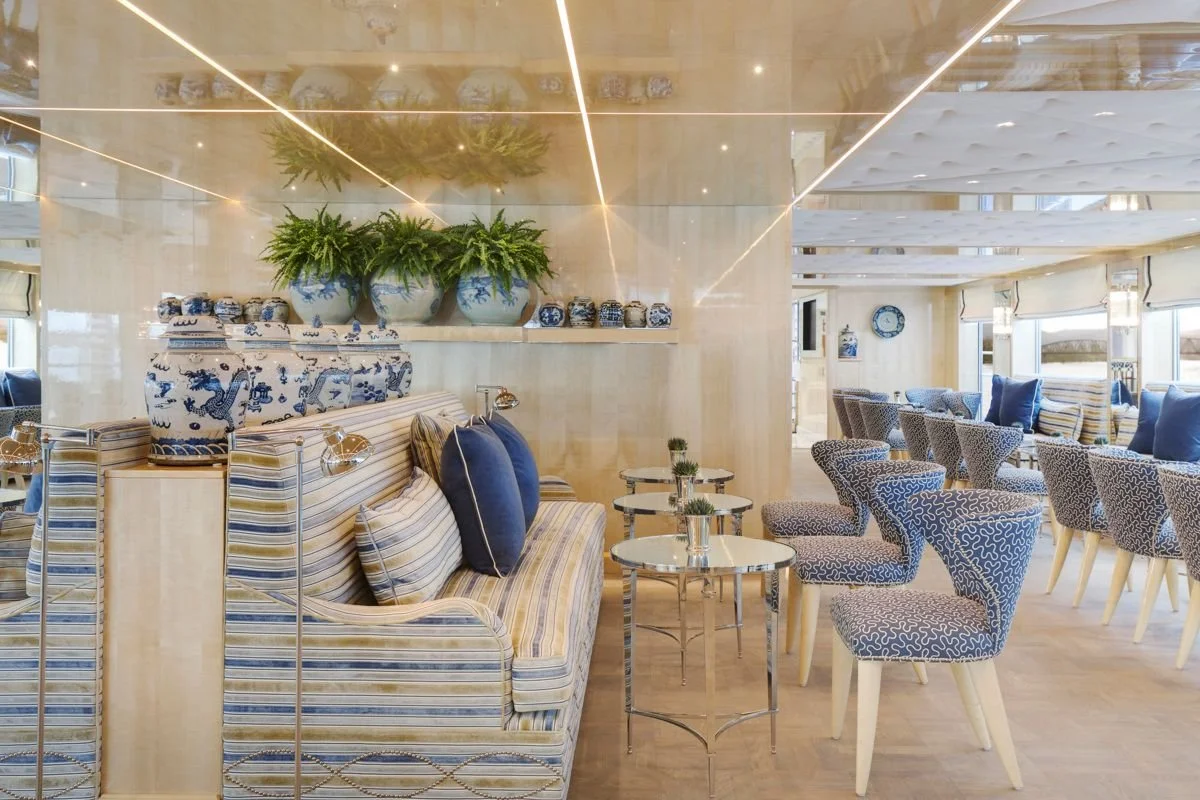 Inside a bright, luxurious restaurant or lounge with a nautical theme, featuring striped sofas with blue and beige cushions, small round tables with potted plants, patterned chairs, and decorative blue and white porcelain jars and vases.