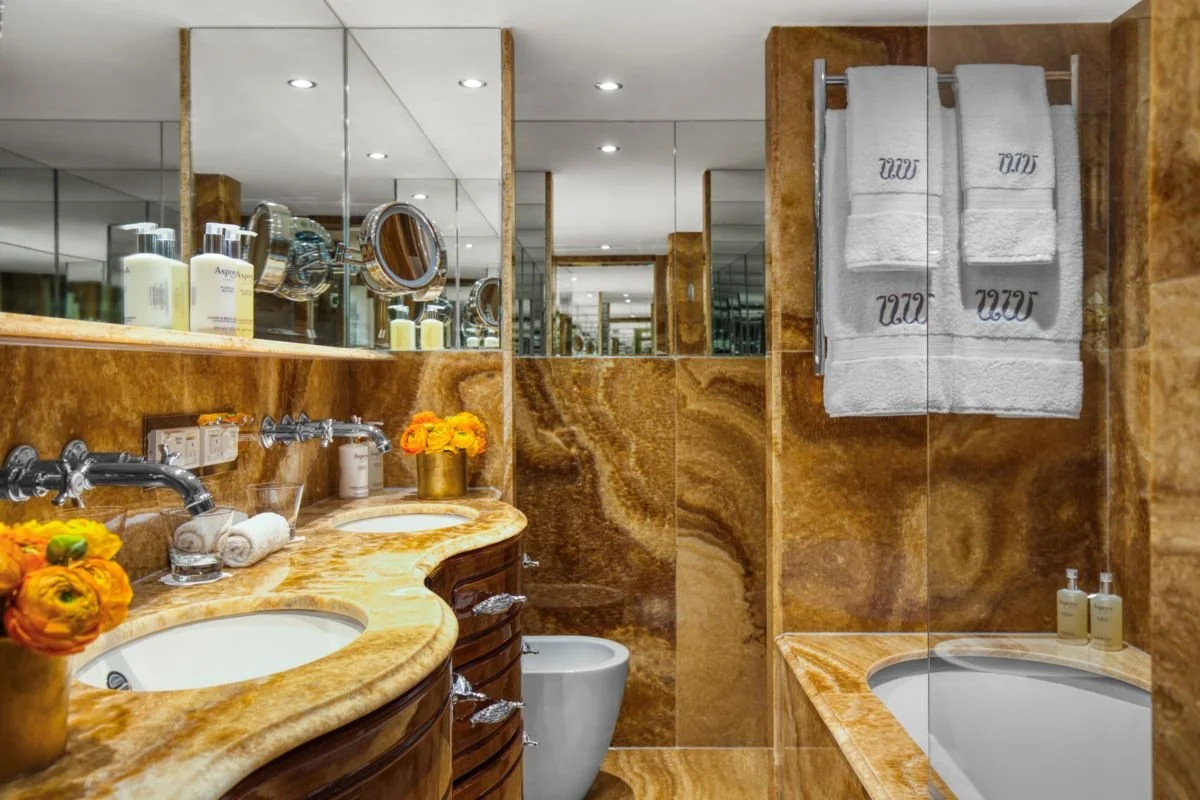 A luxurious bathroom with marble countertops and walls, featuring double sinks with gold vases of orange flowers, a mirror, wall-mounted soap dispensers, towels hanging on a rack, and a bathtub with mounted soap bottles.