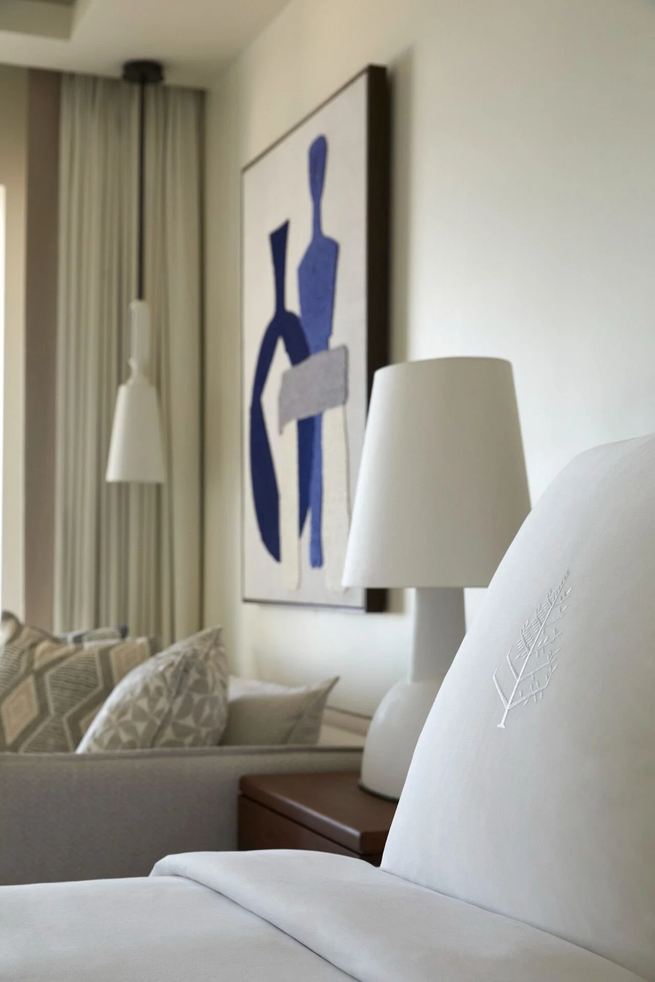 A bedroom with white upholstered furniture, decorative pillows, a wooden side table with a white lampshade, and artwork on the wall depicting a blue figure holding a book or box.