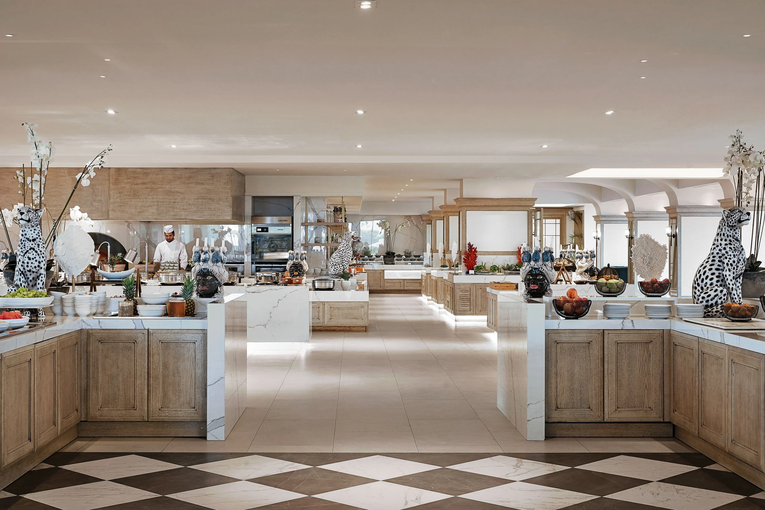 A luxurious buffet setup in a well-lit, elegant kitchen with wooden cabinets, marble countertops, and decorative cheetah and animal masks, with a chef preparing food in the background.