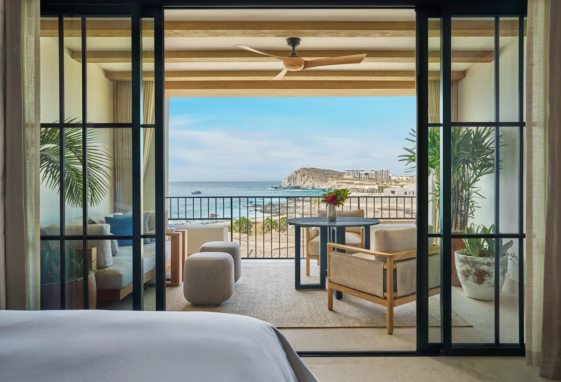 View from a bedroom looking out onto a balcony with an ocean and rocky hill in the background, featuring a sailboat on the water, a small table with a flower arrangement, and cushioned chairs for seating.