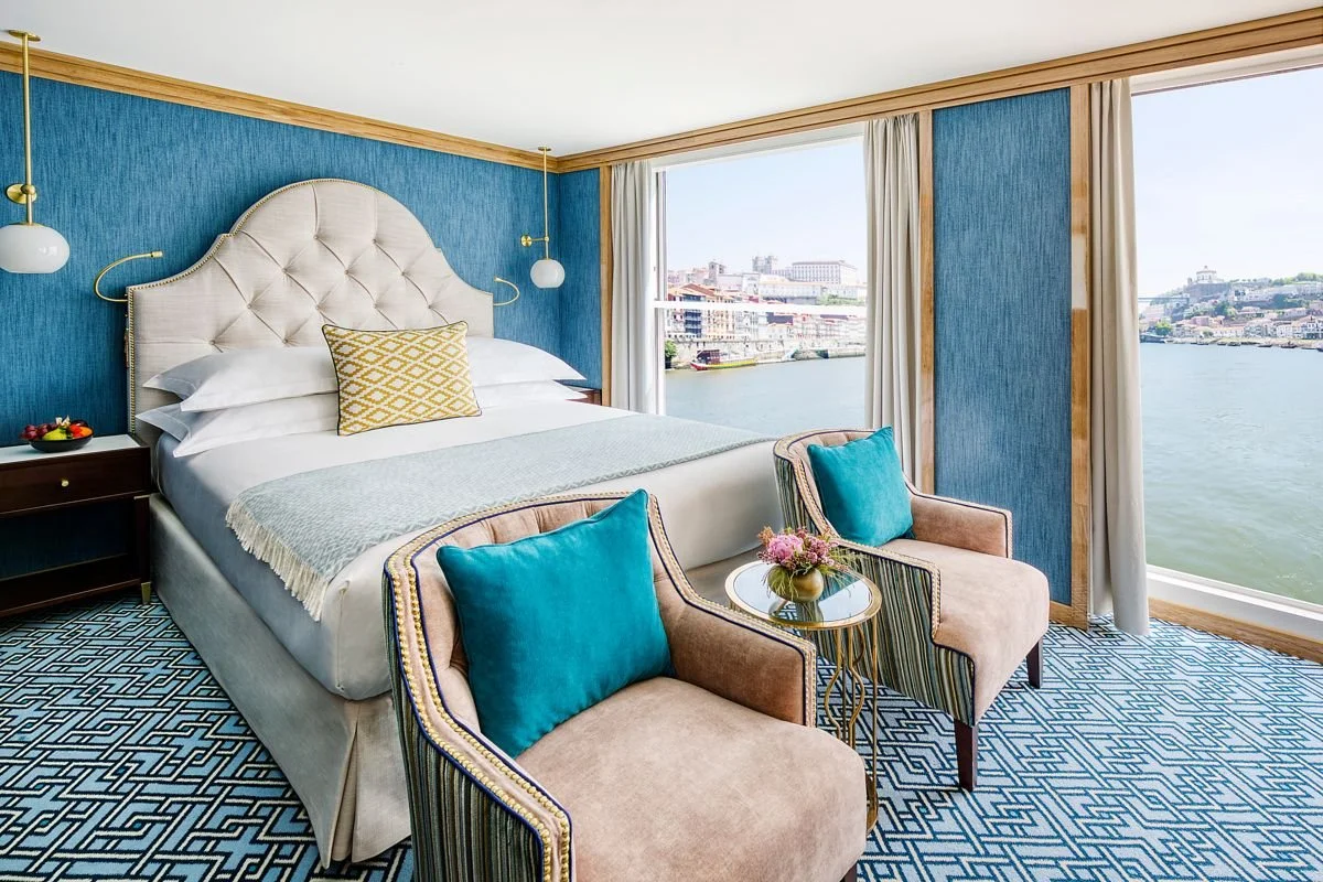 A hotel room with a large bed with a tufted headboard, blue walls, and a view of a river and cityscape through large windows.