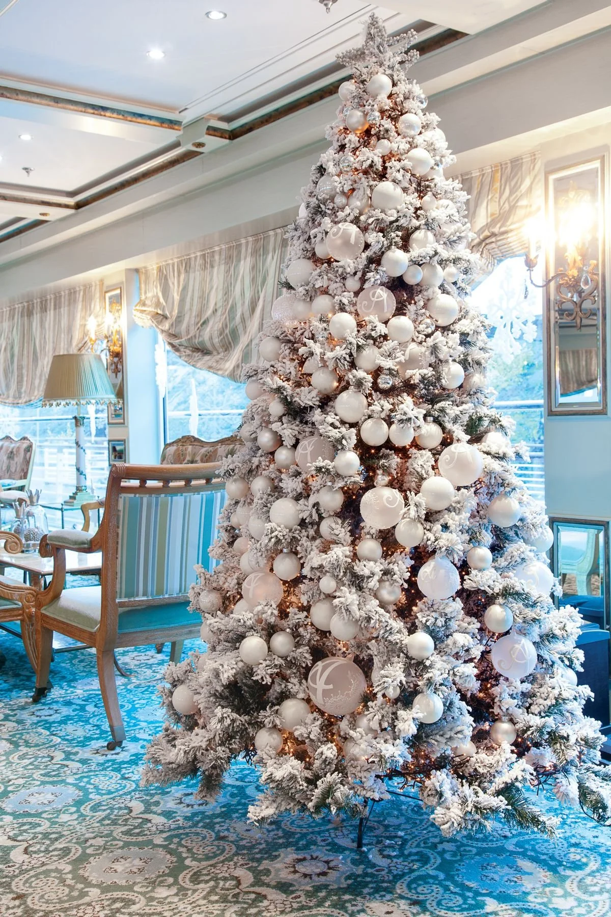 A decorated Christmas tree covered in artificial snow, adorned with white and silver ornaments, in a well-lit, ornate living room with vintage furniture and large windows.