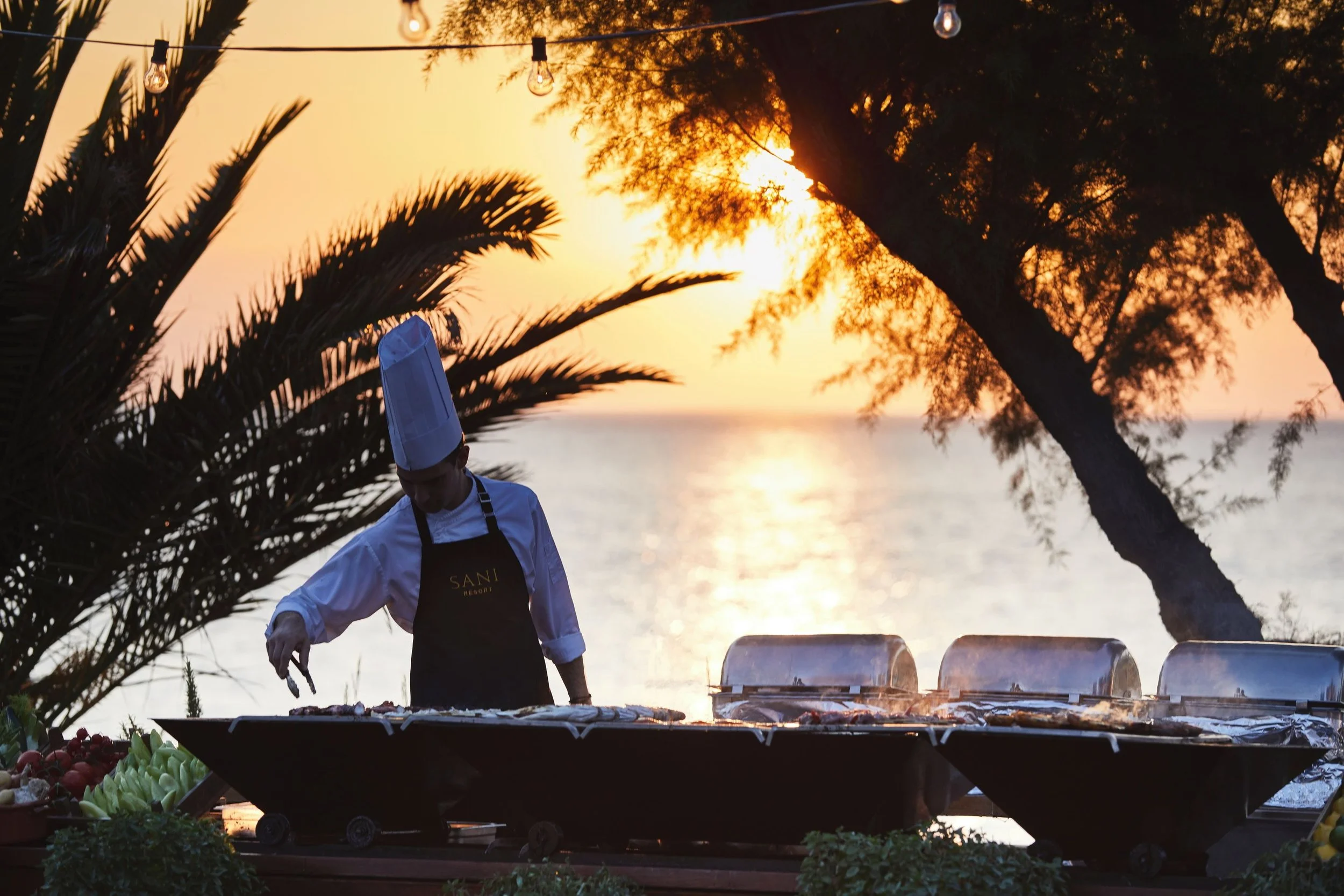 A chef wearing a white uniform and chef's hat preparing food outdoors during sunset, with string lights hanging above and trees in the background.