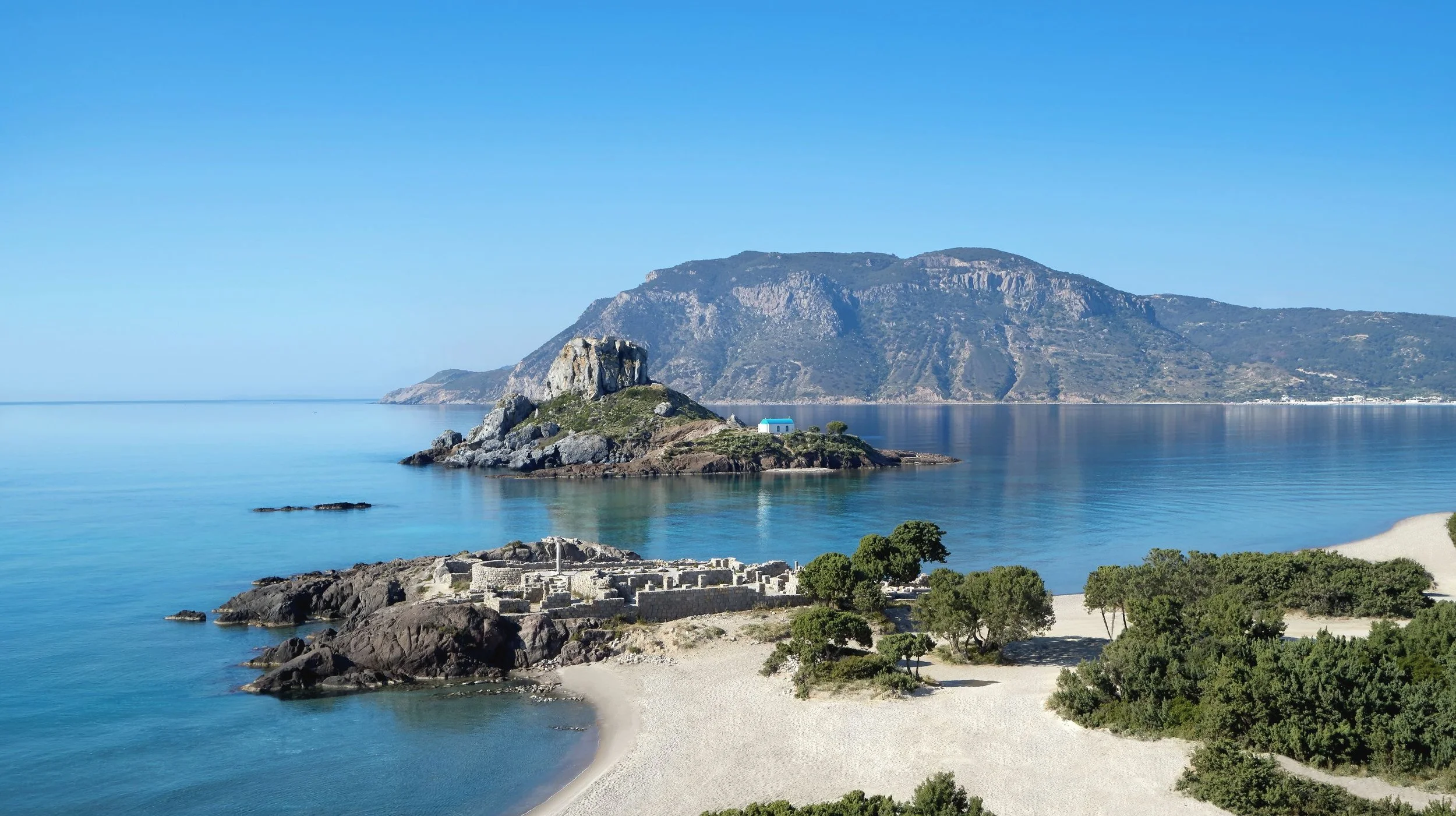 A coastal landscape with a small island featuring a white building with a blue roof, rocky shoreline, green trees, and a large mountain in the background under a clear blue sky.