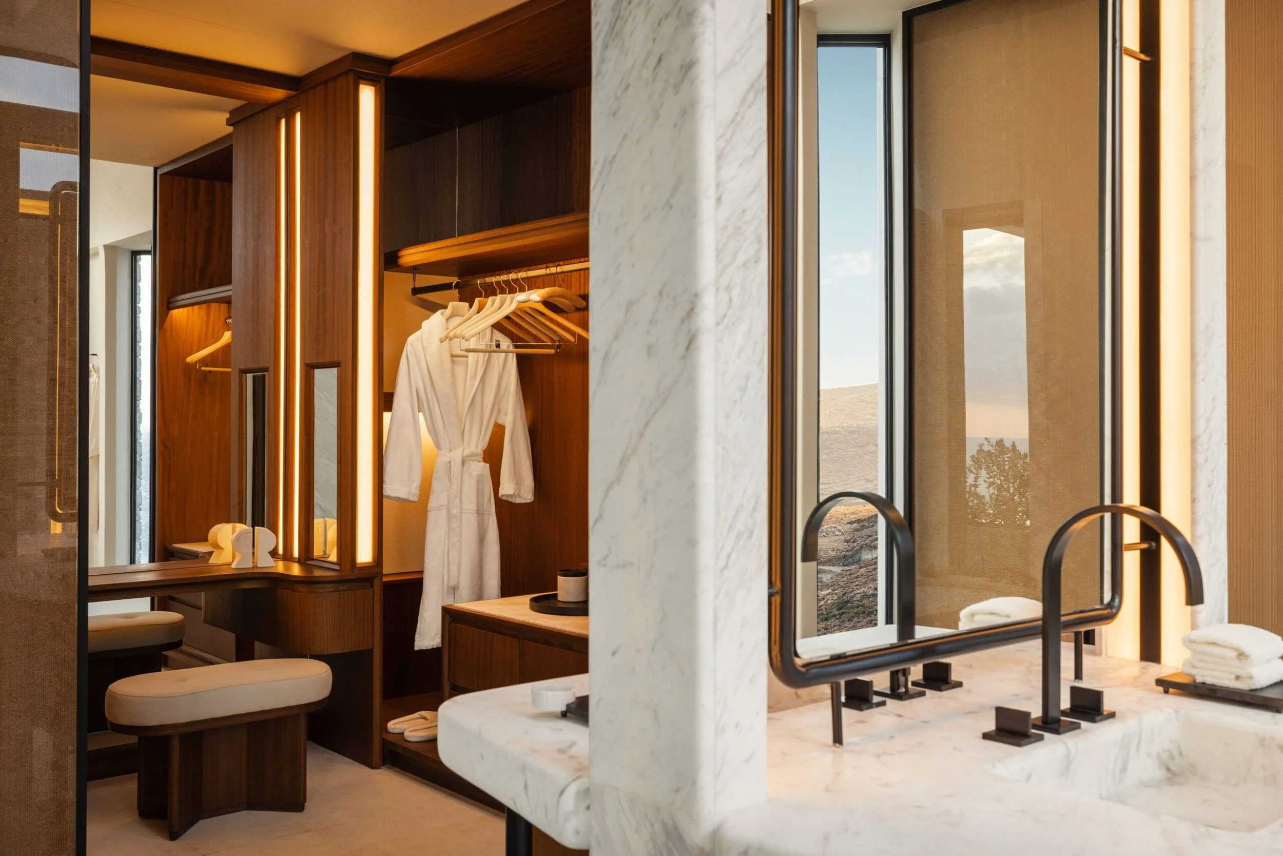 Modern bathroom with marble countertops, black fixtures, a window revealing a landscape, and a wooden wardrobe with hanging robes and towels.