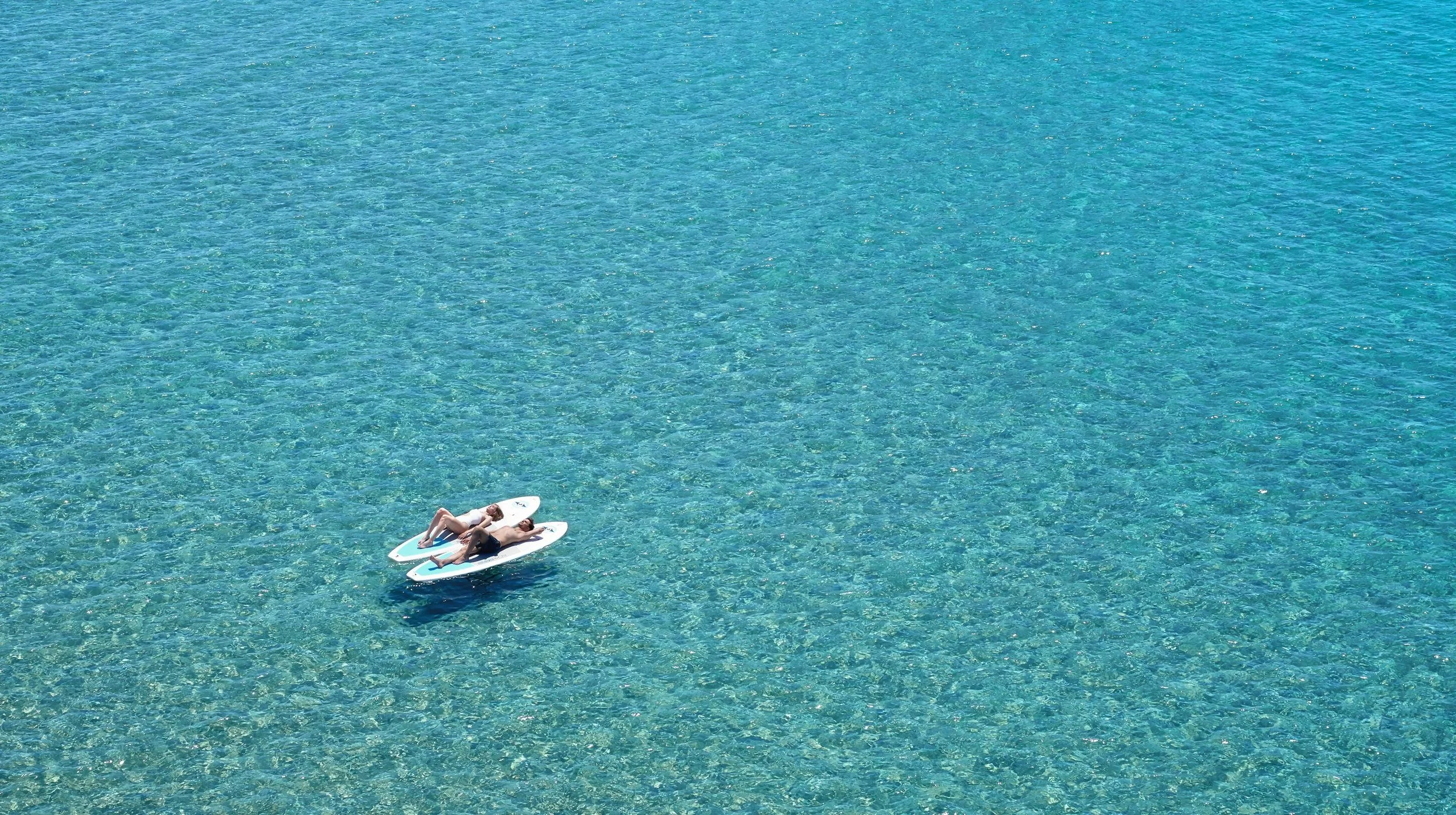Two people are lying on paddleboards in clear blue water, relaxing and sunbathing.