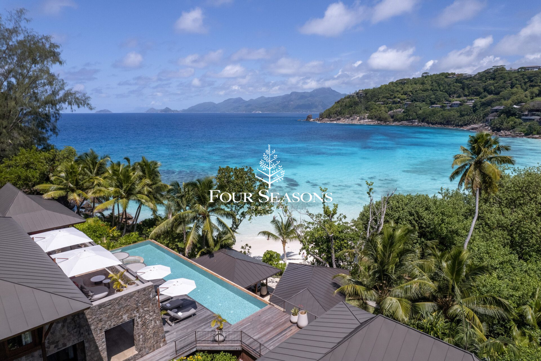 Aerial view of Four Seasons Resort with pool area, beach, palm trees, ocean, and mountains in the background.