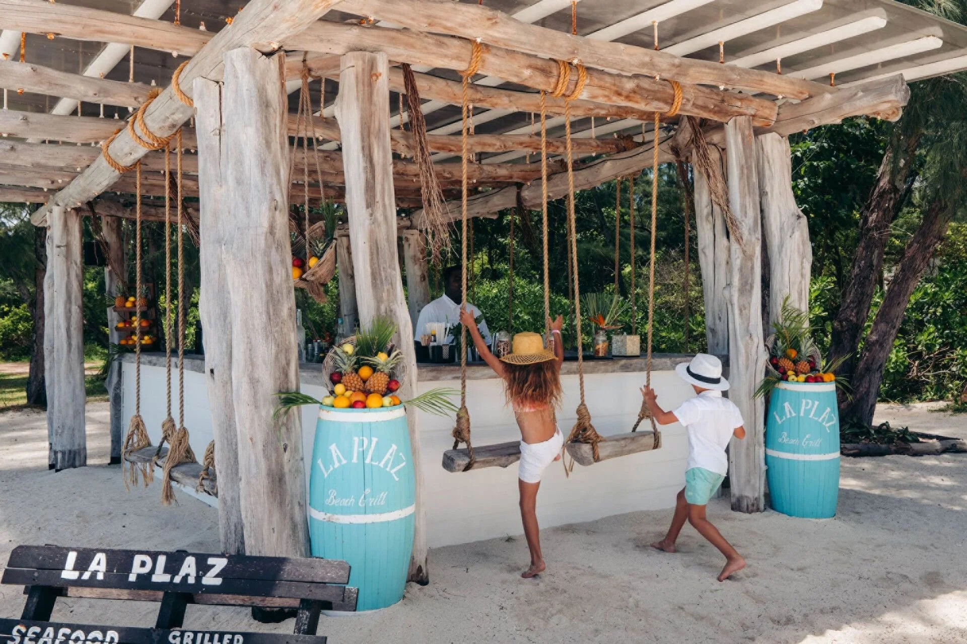 A beachside bar with a rustic wooden structure and swings hanging from the roof. Two children are playing near the bar, and a person behind the bar is preparing drinks. There are large blue barrels with fruit decorations and signs that read 'La Plaz Beach Grill' and 'Seafood'. The area is surrounded by greenery and sand.