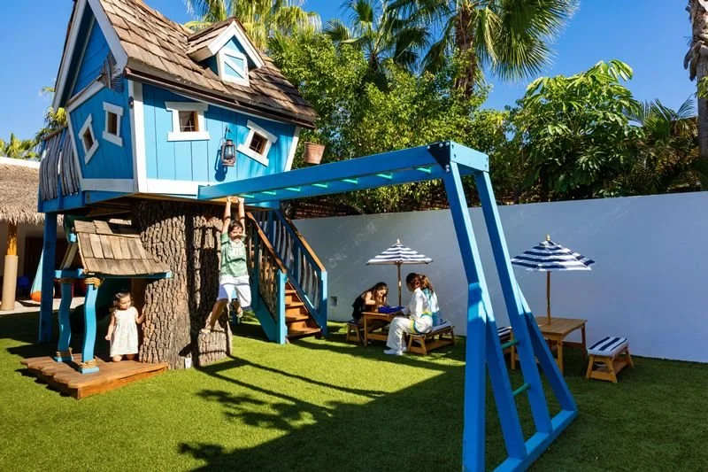 Children playing outside in a backyard with a treehouse, picnic table with adults, and pools with umbrellas, surrounded by lush greenery and a white fence.
