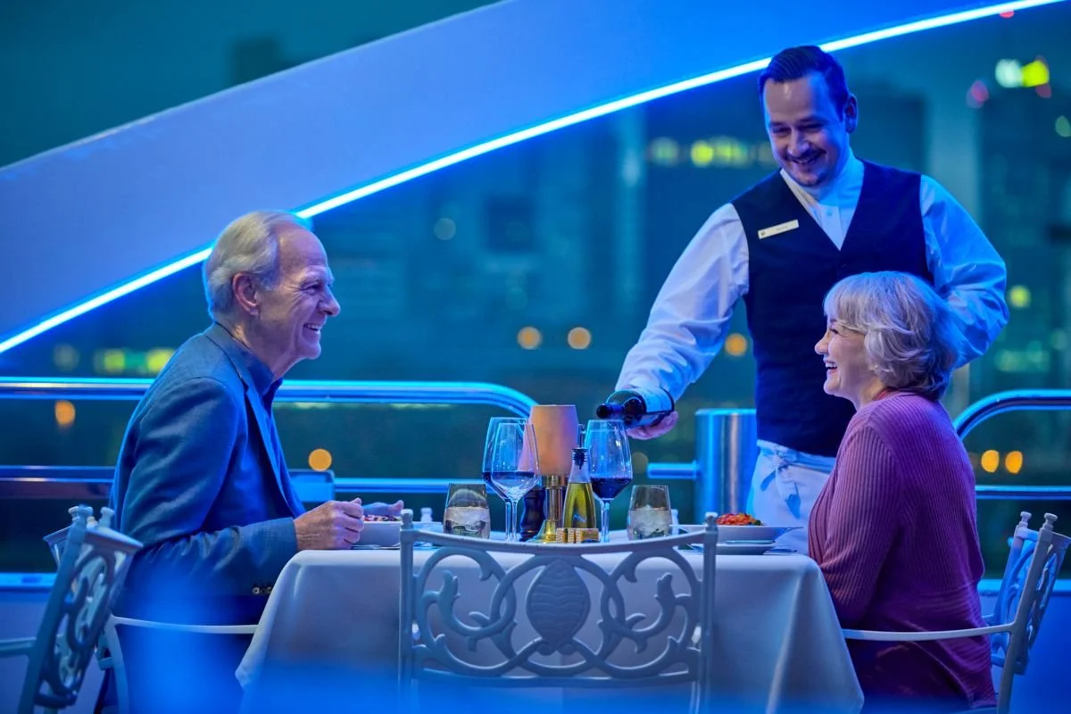 A waiter pouring wine for a smiling elderly couple dining at a table in a modern restaurant with blue neon lighting.