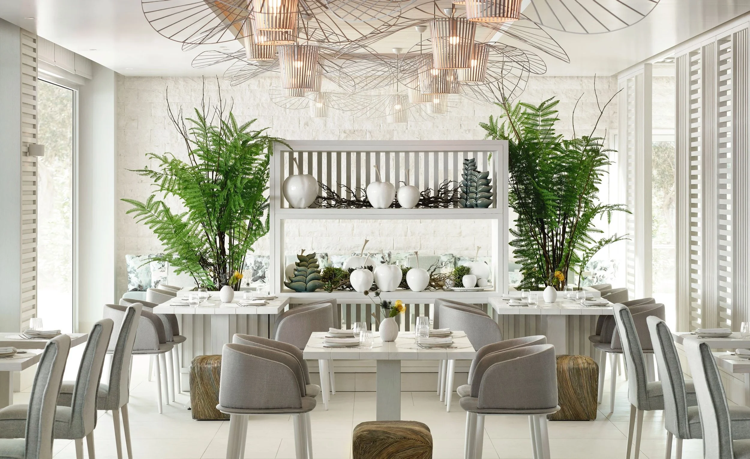 Elegant dining room with large windows, white walls, and decorative lighting. Tables are set with white plates, glasses, and small vases with flowers. Green plants and modern white shelves with decorative items add to the stylish decor.
