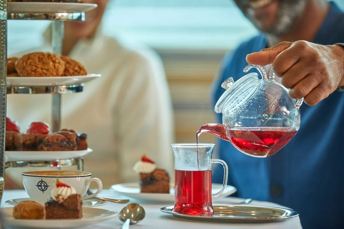 Person pouring red tea from a glass teapot into a glass cup on a table with desserts and a tiered tray of pastries in the background.