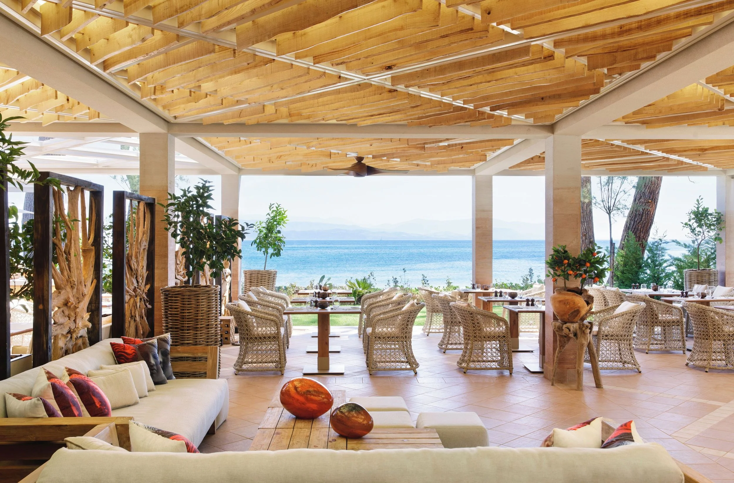 Open-air restaurant with wooden ceiling and beach view, wicker chairs, and outdoor plants.