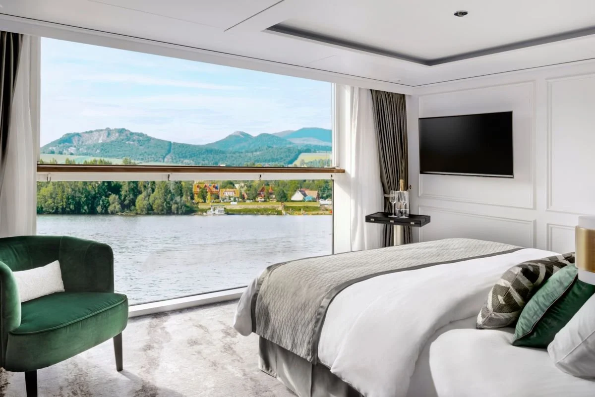 Luxury hotel room with a large window overlooking a river, trees, and mountains in the distance. The room has a white bed with decorative pillows, a green armchair, a small black table with glass water pitchers, and a flat-screen TV on the wall.