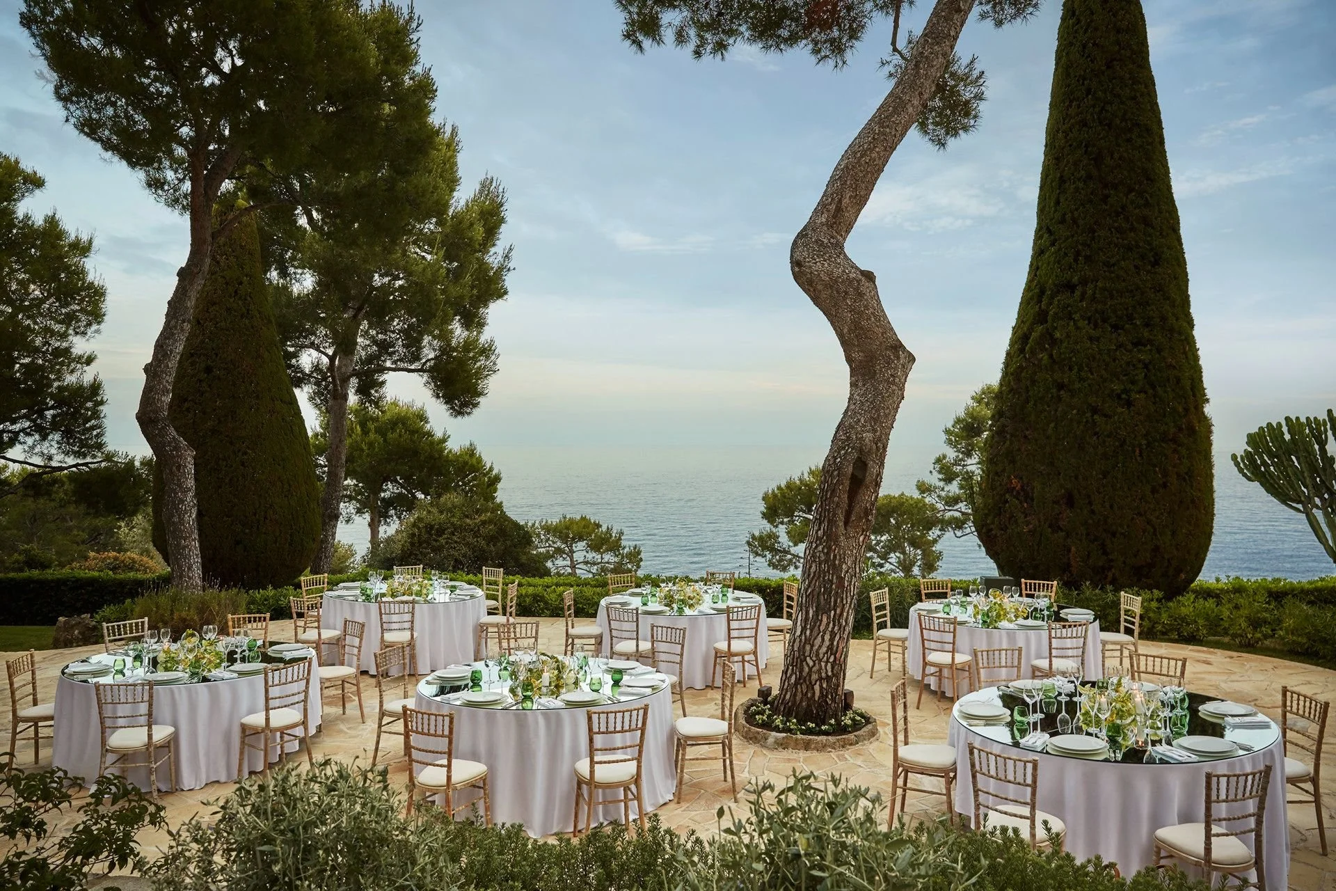 Outdoor dining setup with round tables covered in white tablecloths, surrounded by wooden chairs, on a patio overlooking the ocean. Tall trees and lush greenery frame the scene, creating a scenic, elegant setting.