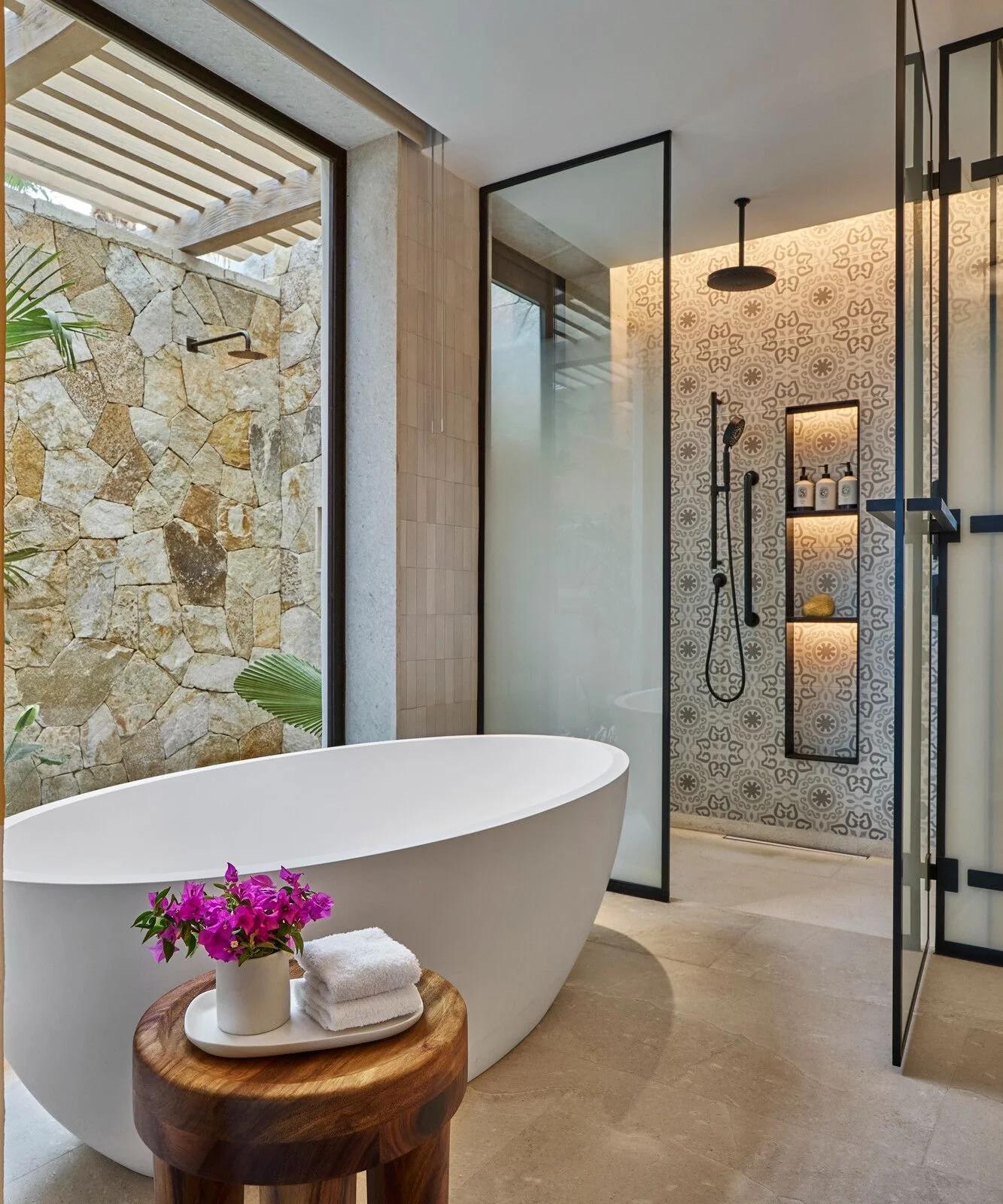 Modern bathroom with a freestanding bathtub near a window with a stone wall outside. The shower area has patterned tiles, a black showerhead, and a glass partition. There are plants and a small wooden table with towels and purple flowers.
