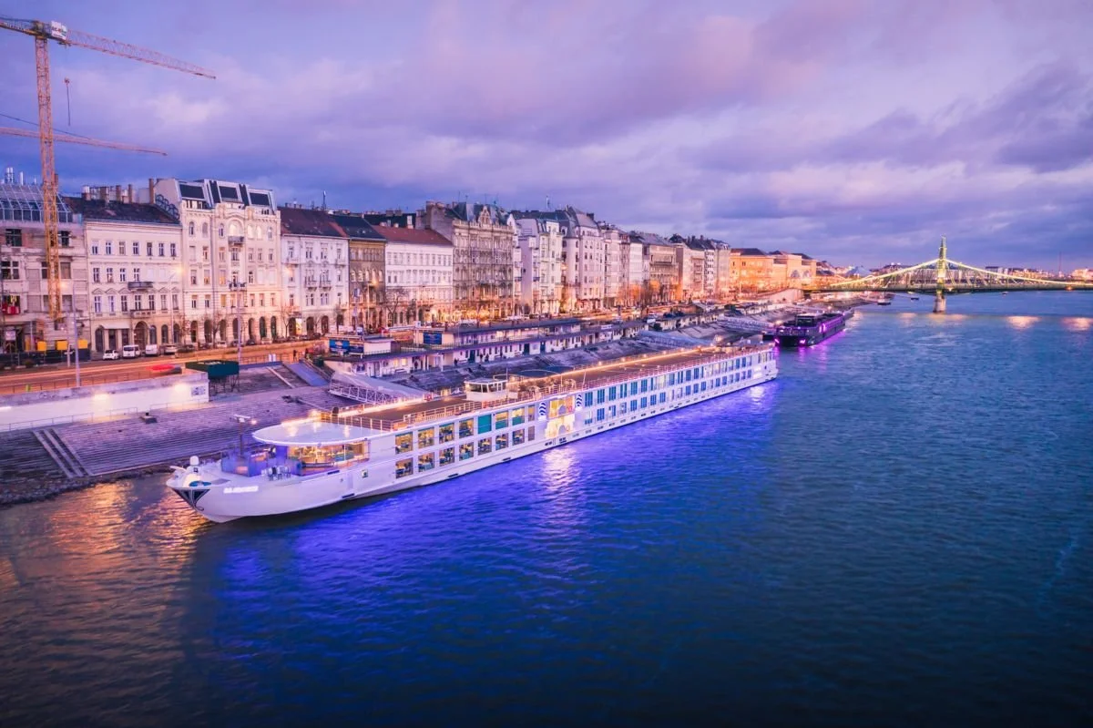 A river with a large white cruise ship docked along the city waterfront during evening twilight, with historic buildings and a bridge illuminated in the background.