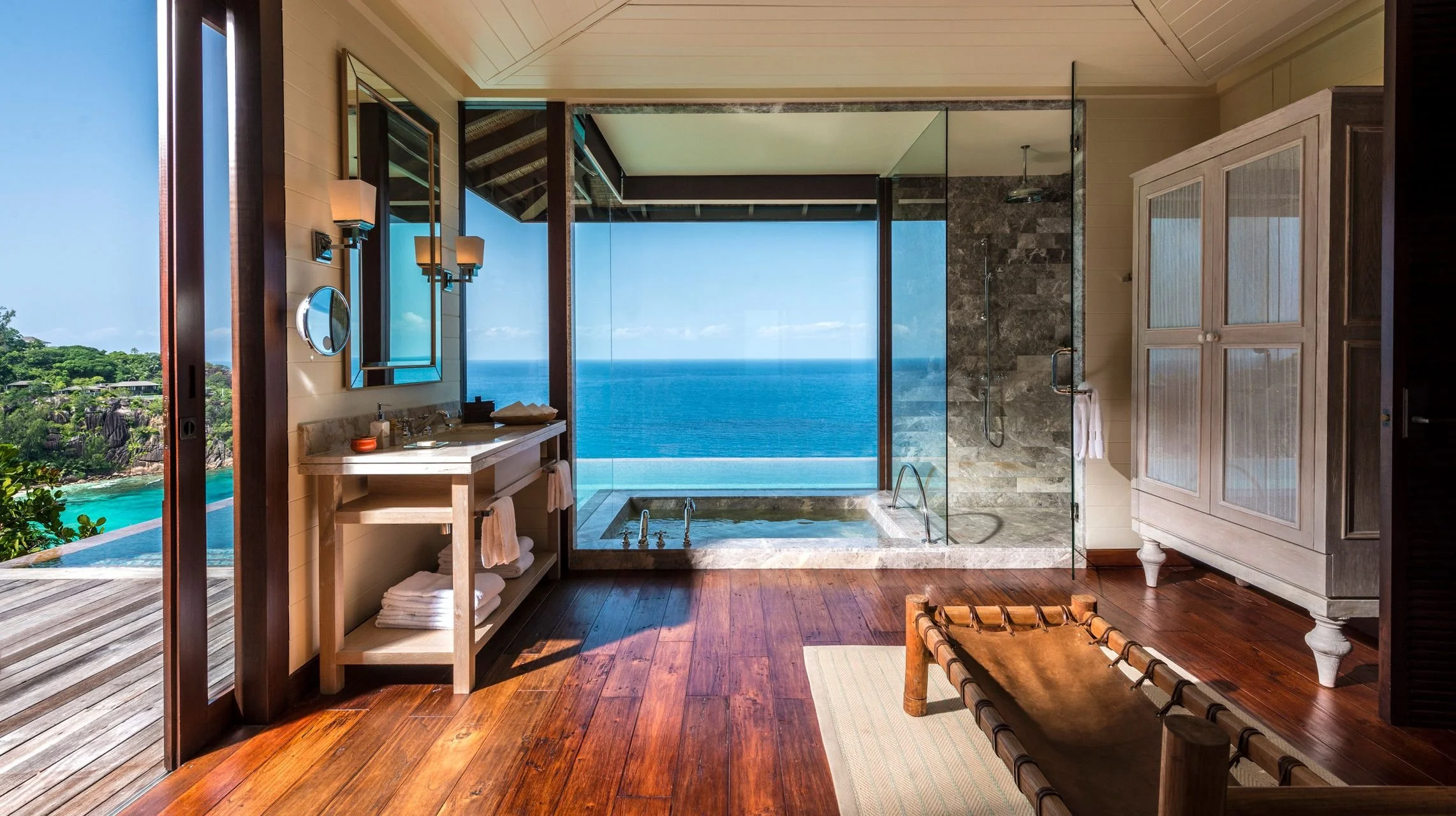A luxurious bathroom with a view of the ocean through a large glass window, featuring a stone shower area, a vanity with toiletries, and a wooden bench, with wooden flooring and soft lighting.