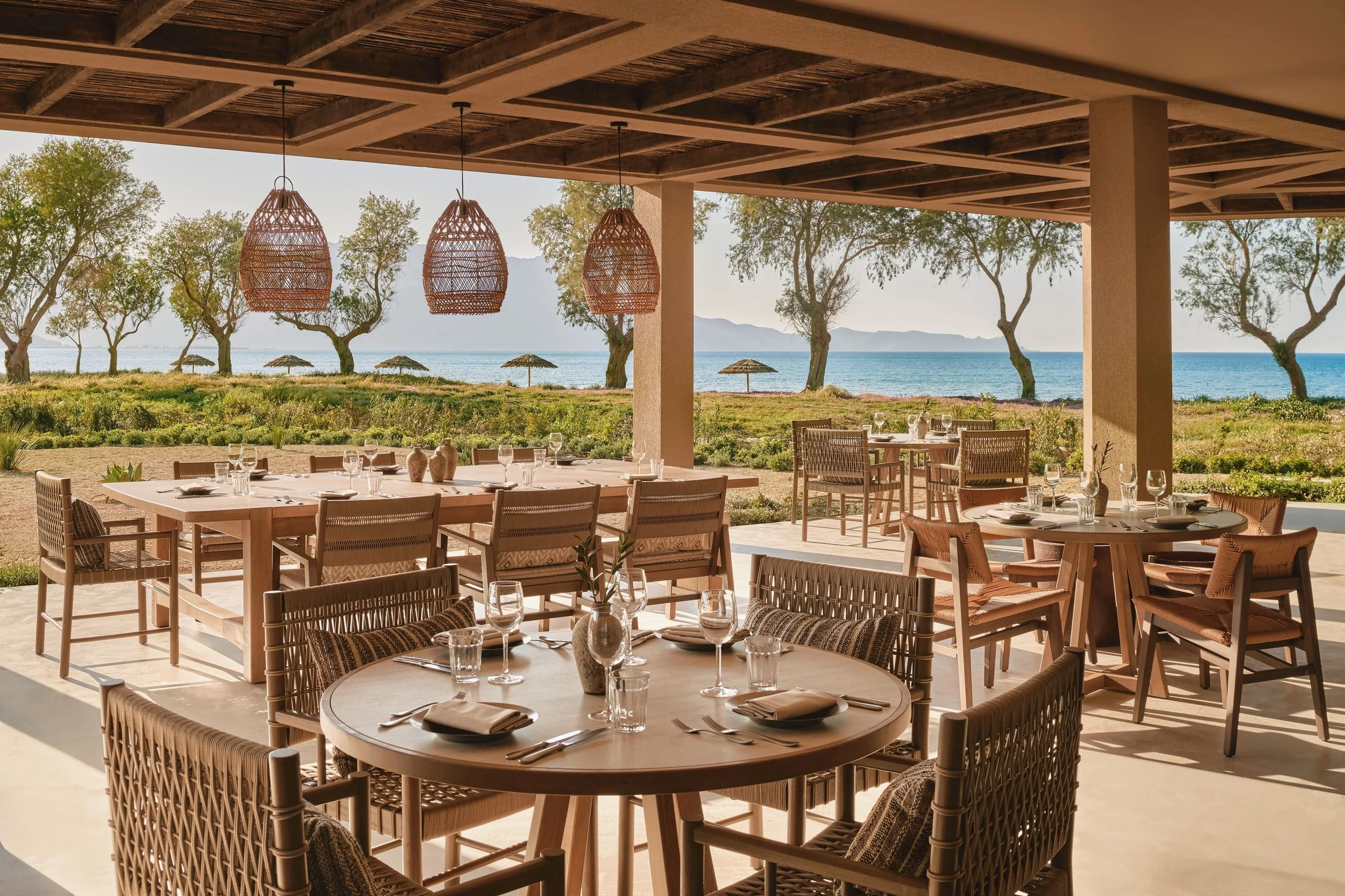 Outdoor restaurant seating area with round and rectangular tables, wicker chairs, and table settings near a beach with trees and umbrellas