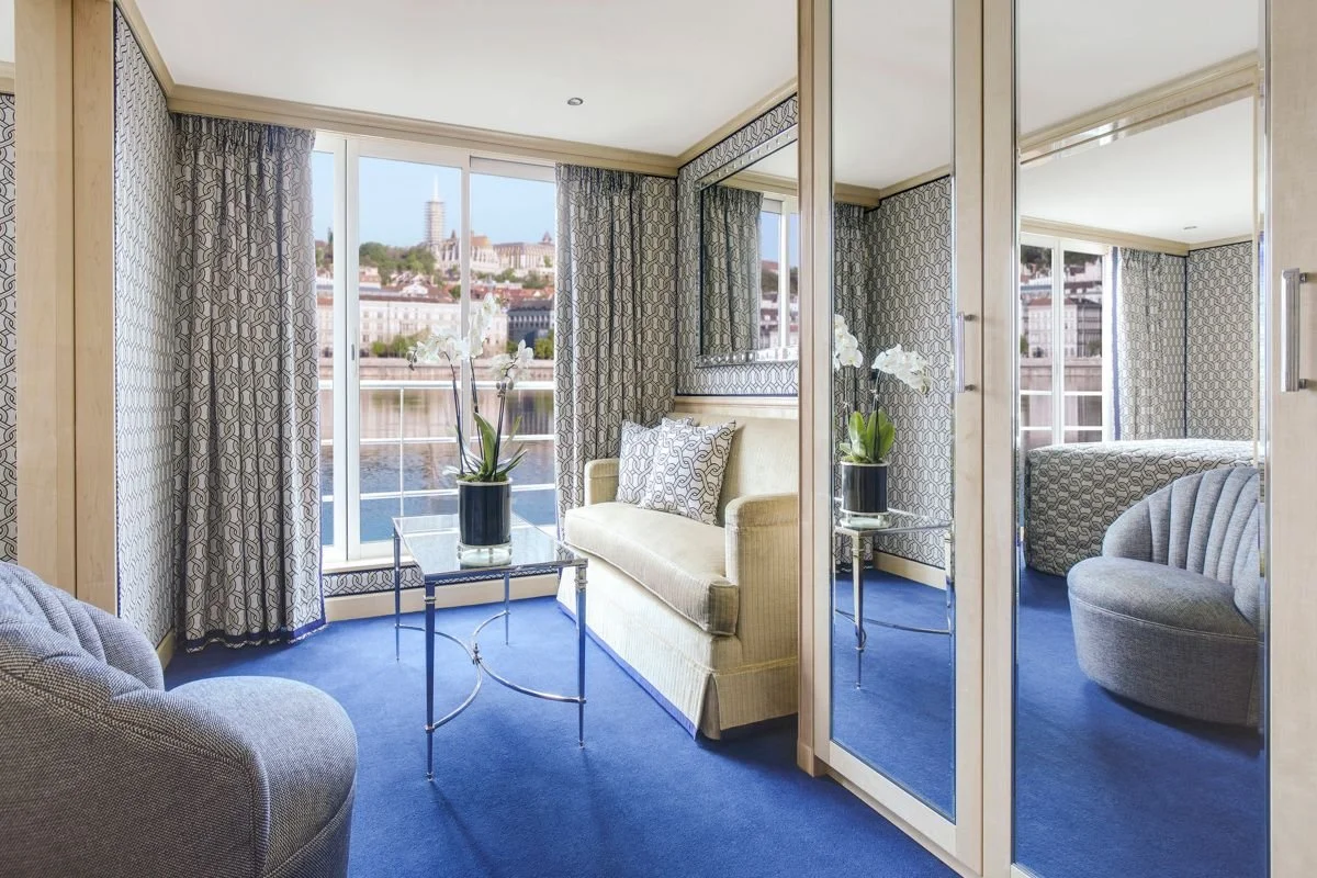 Living room with a cream sofa and a gray armchair, a glass coffee table with a potted orchid, and a large window with curtains overlooking a cityscape.