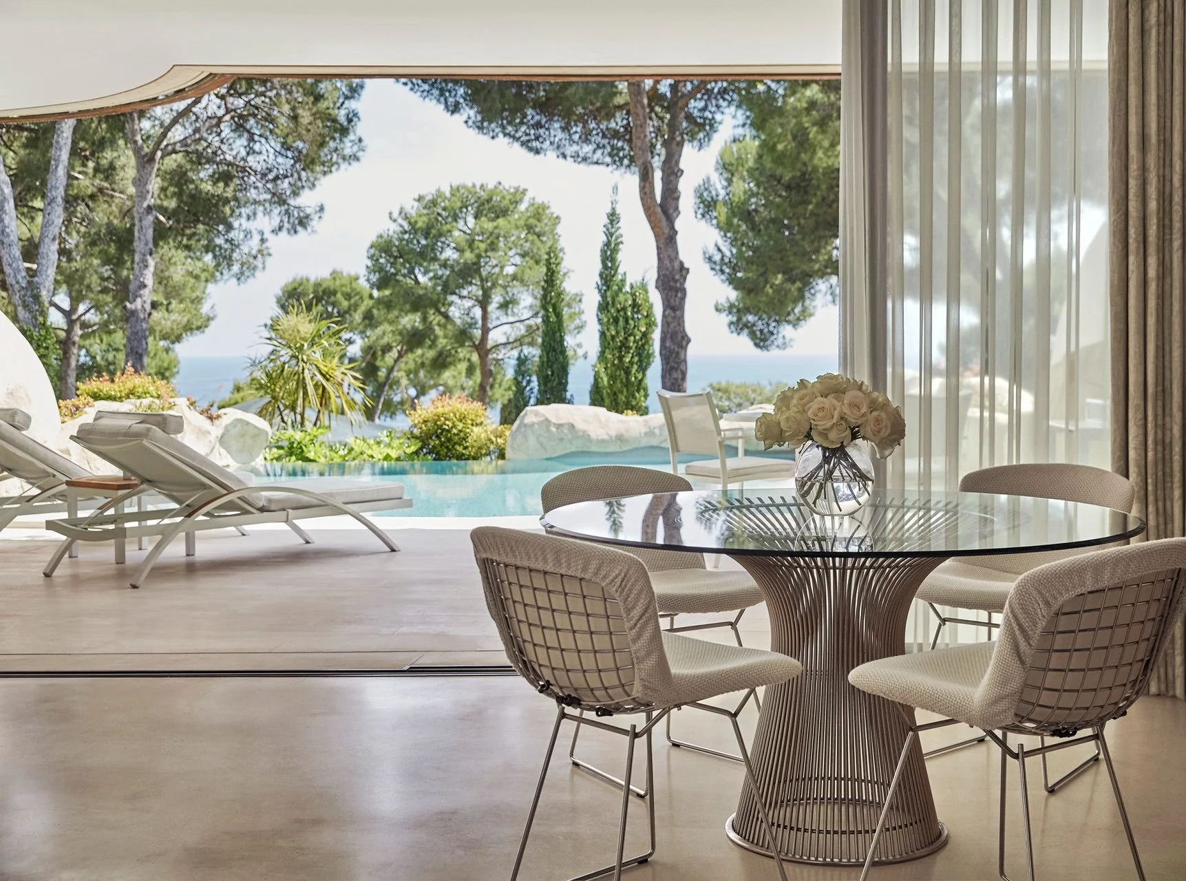 Elegant dining area with a glass-top round table and beige chairs, decorated with a bouquet of white roses, overlooking an outdoor pool and scenic trees.