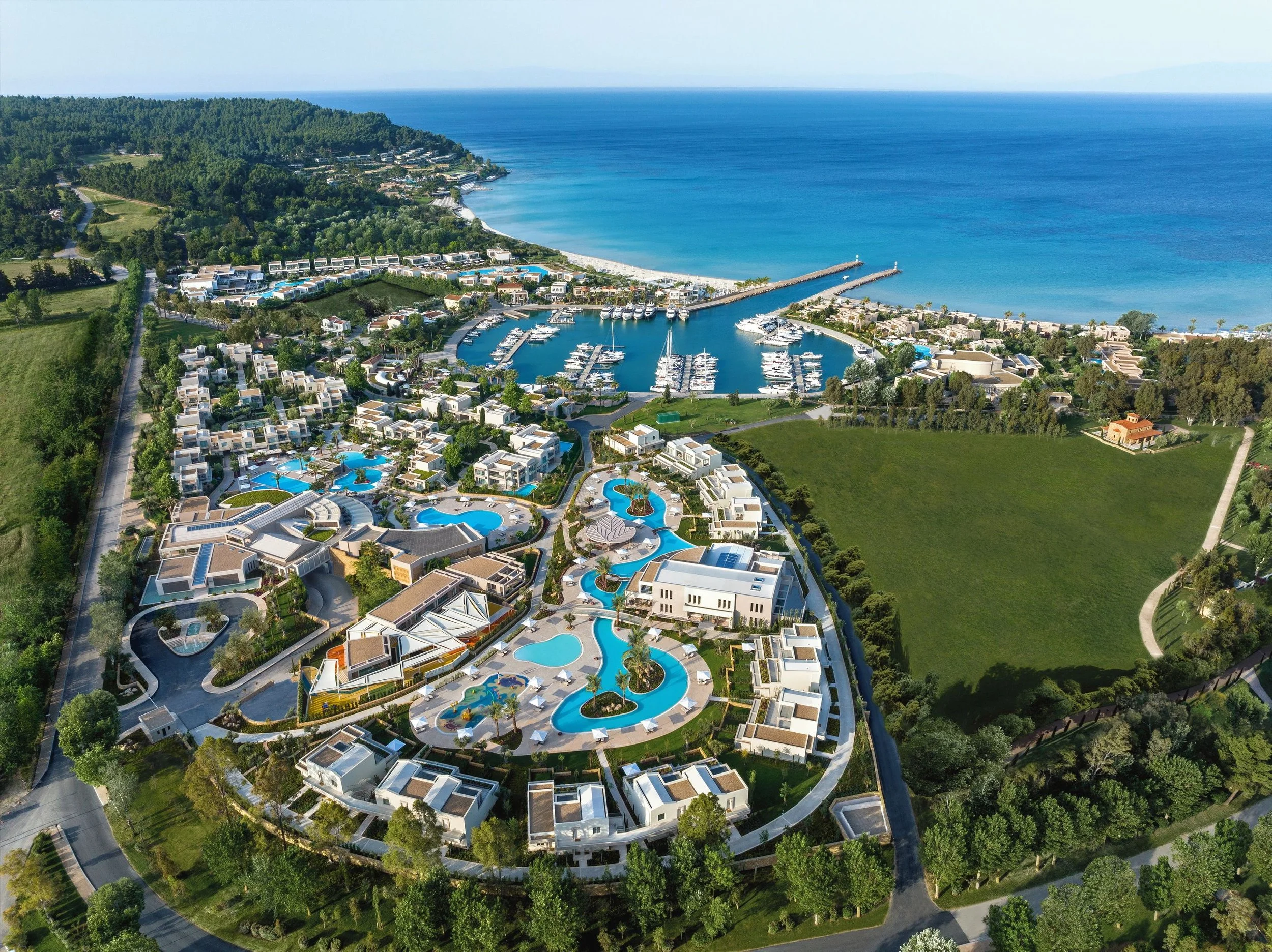 Aerial view of a coastal resort with white buildings, swimming pools, and a marina with yachts, set against green hills and the ocean.
