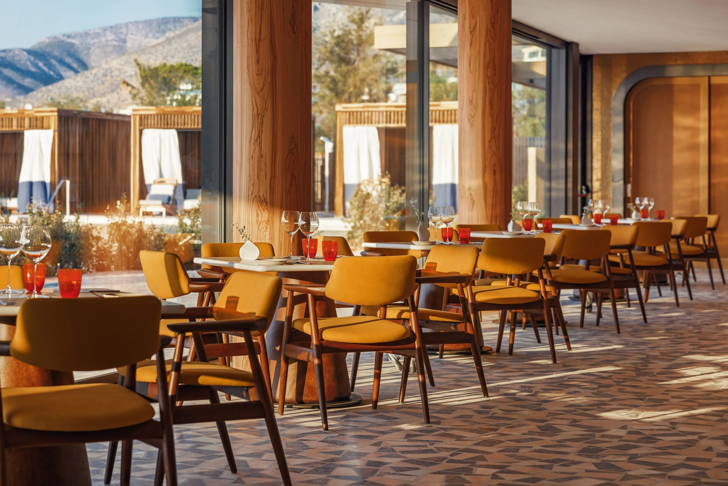 Interior of a modern restaurant with yellow chairs and white tables set with glassware and candles, large windows with wooden frames reveal an outdoor patio with umbrellas and lounge chairs, mountains are visible in the background.